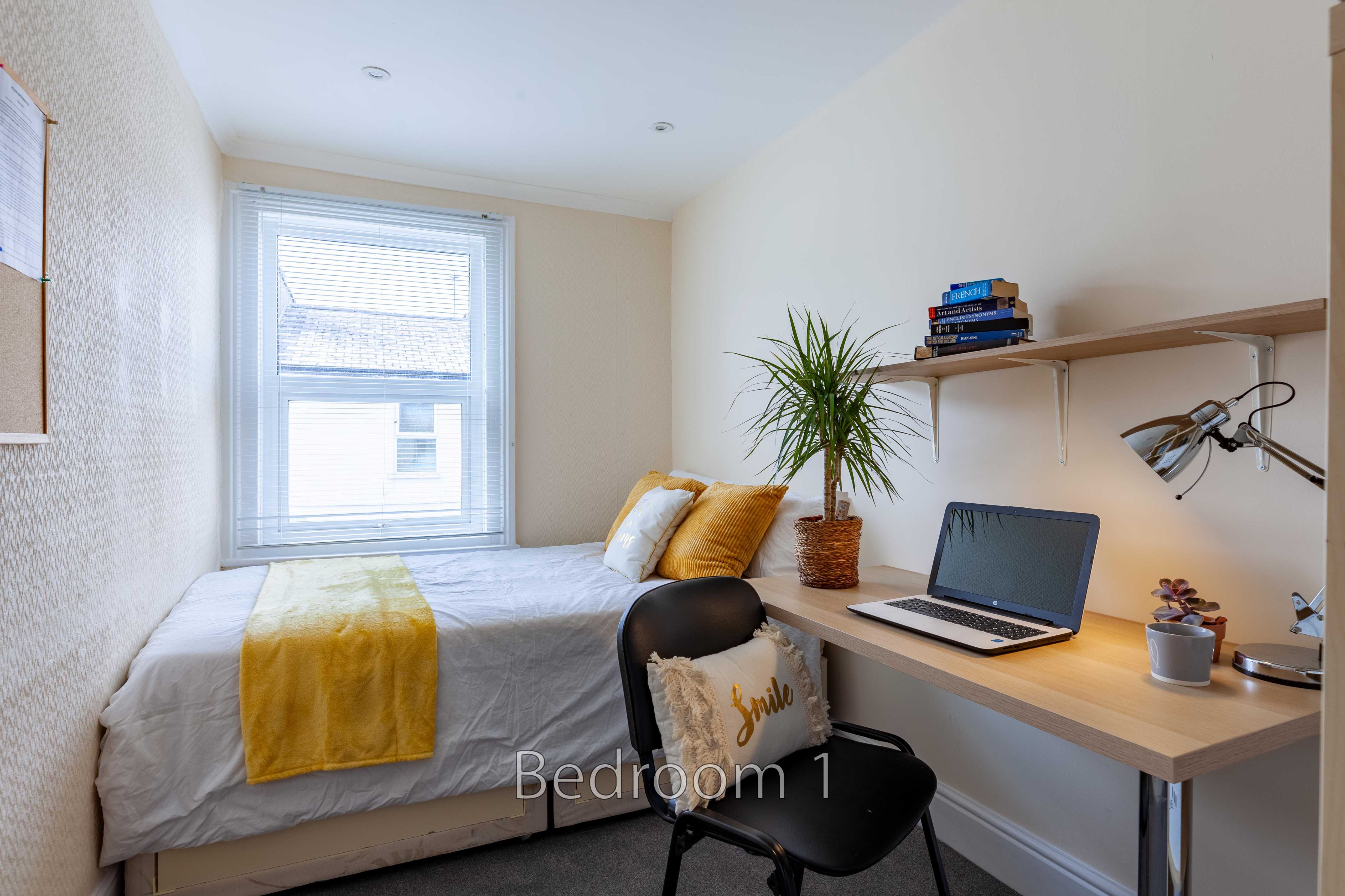 A furnished bedroom with a single bed, desk, chair, shelves and a window.