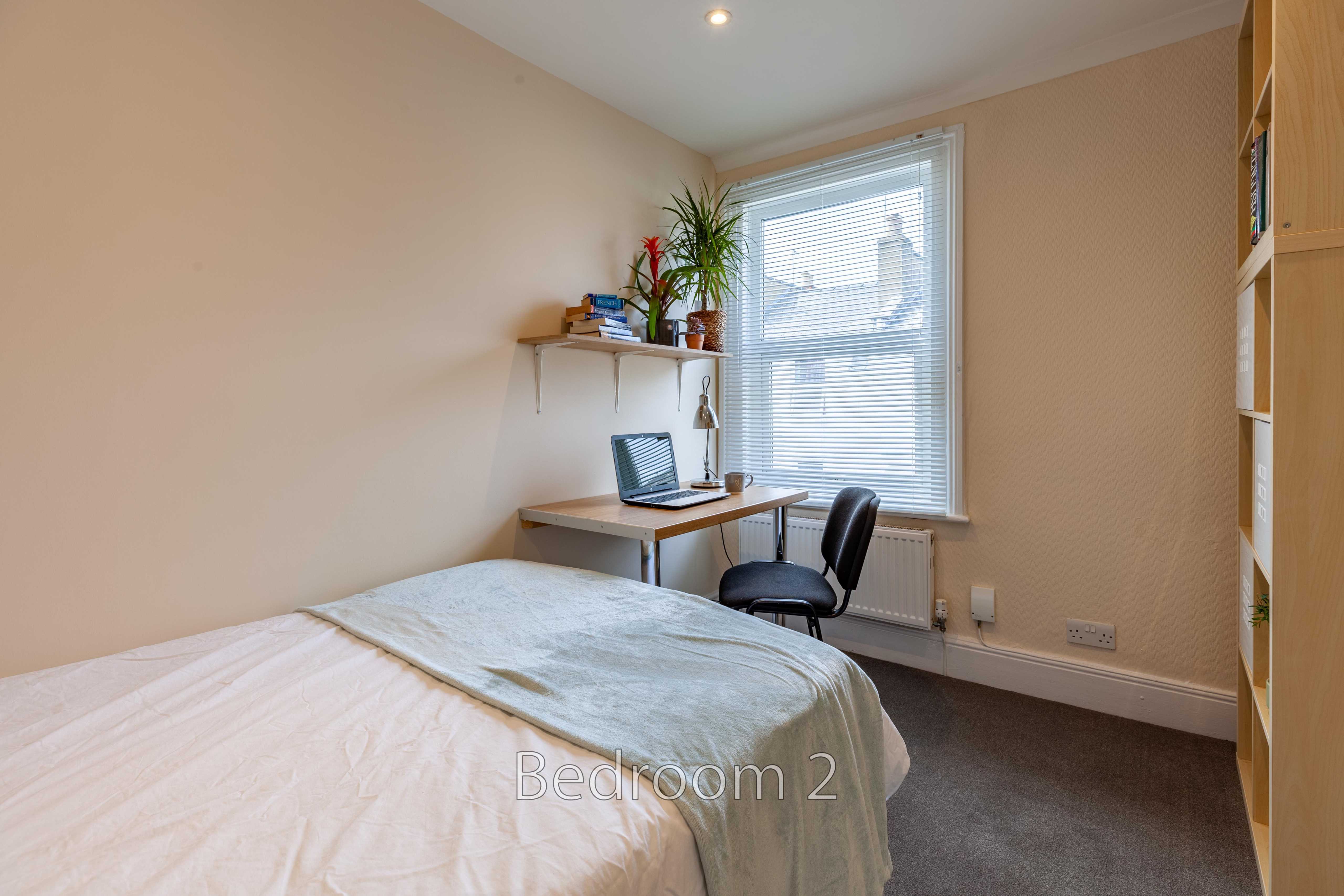 A bedroom with a bed, desk and chair, shelving, a window with blinds, and a radiator.