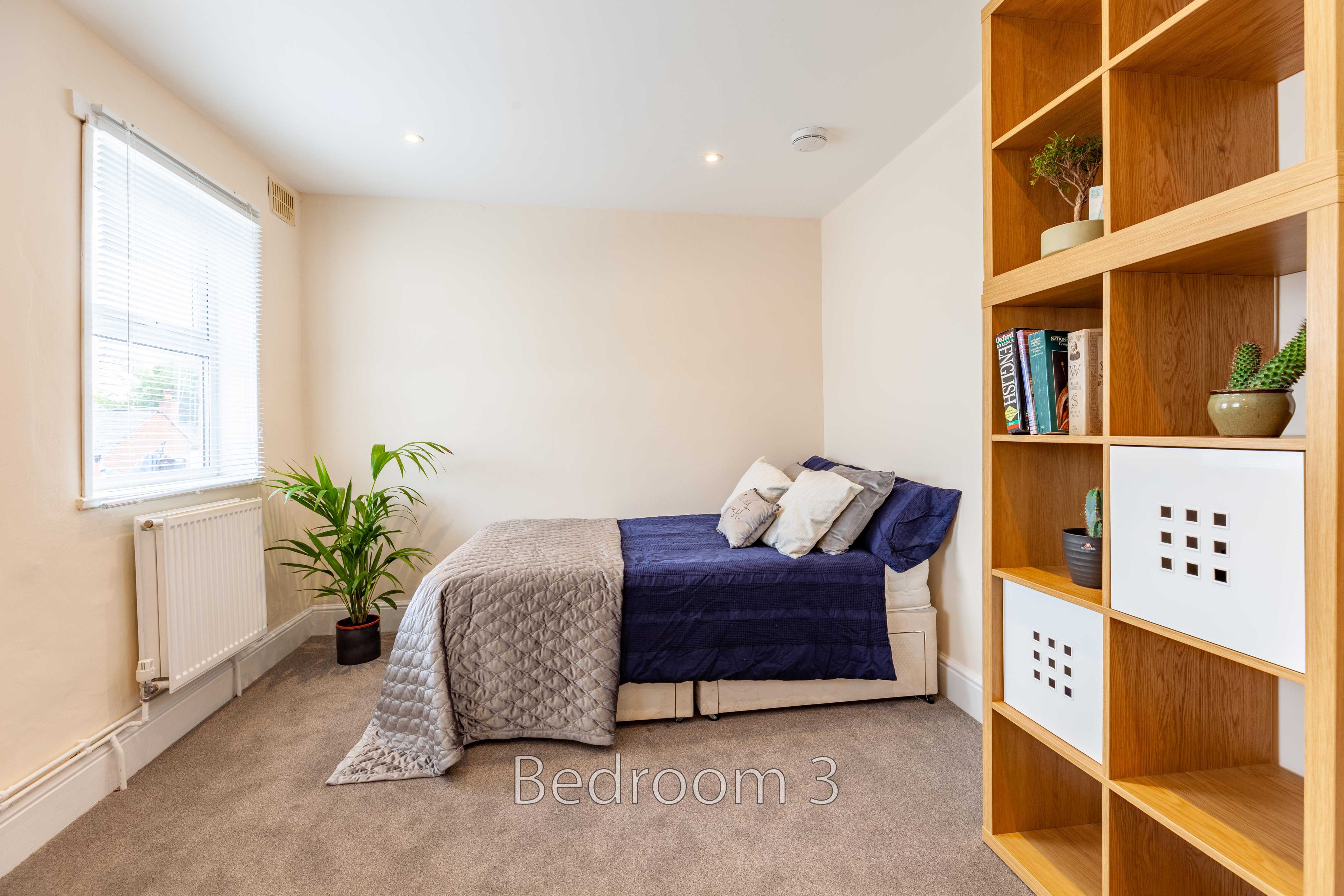 A furnished bedroom with a bed, window, radiator, plant and wooden shelving.