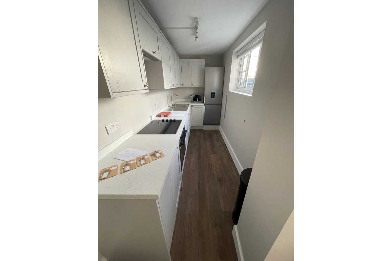 Narrow modern kitchen with white cabinets, countertop, appliances, and dark wood flooring.