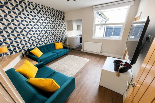 Living room with two teal sofas, yellow cushions, patterned wallpaper, a television and a kitchen area.