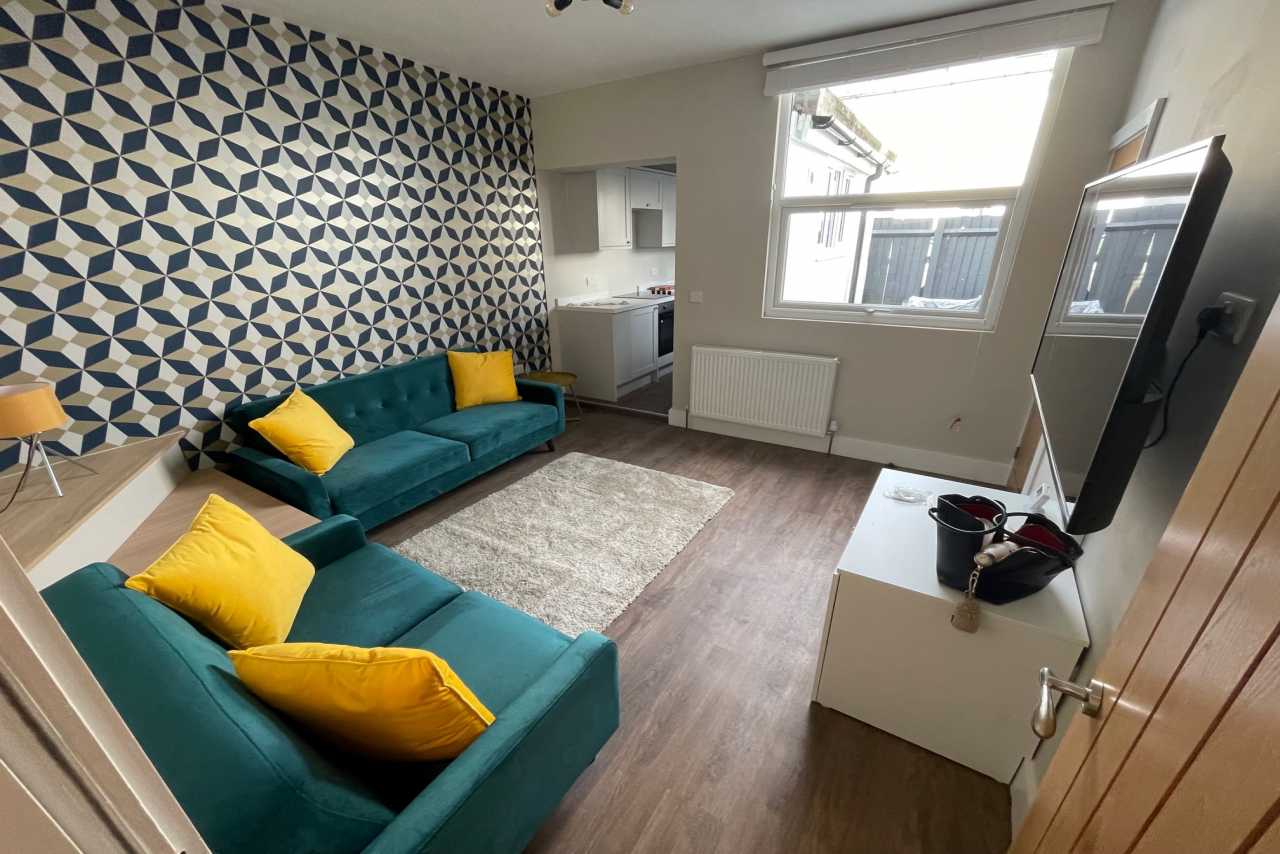 Living room with two teal sofas, yellow cushions, patterned wallpaper, a television and a kitchen area.
