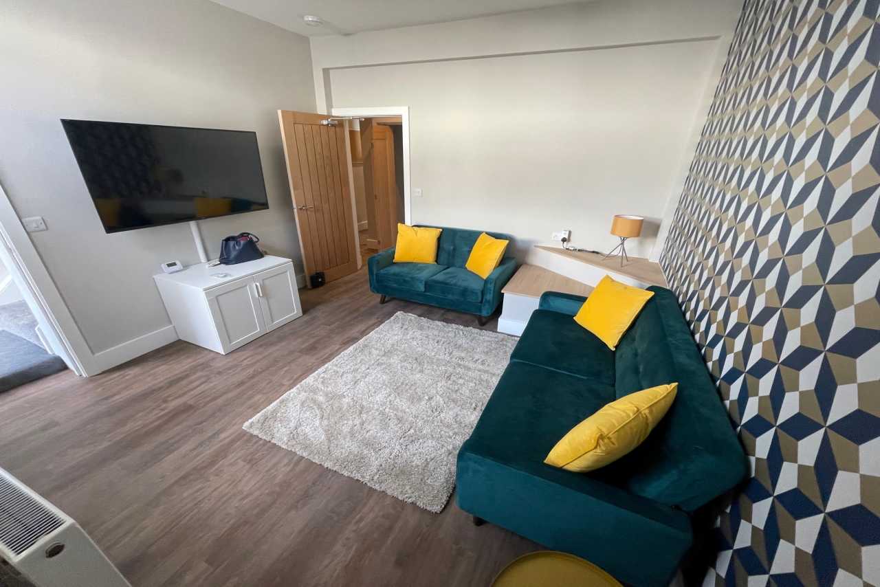 A furnished living room with two teal sofas, yellow cushions, a wall-mounted television, a white media cabinet, a rug and patterned wallpaper.