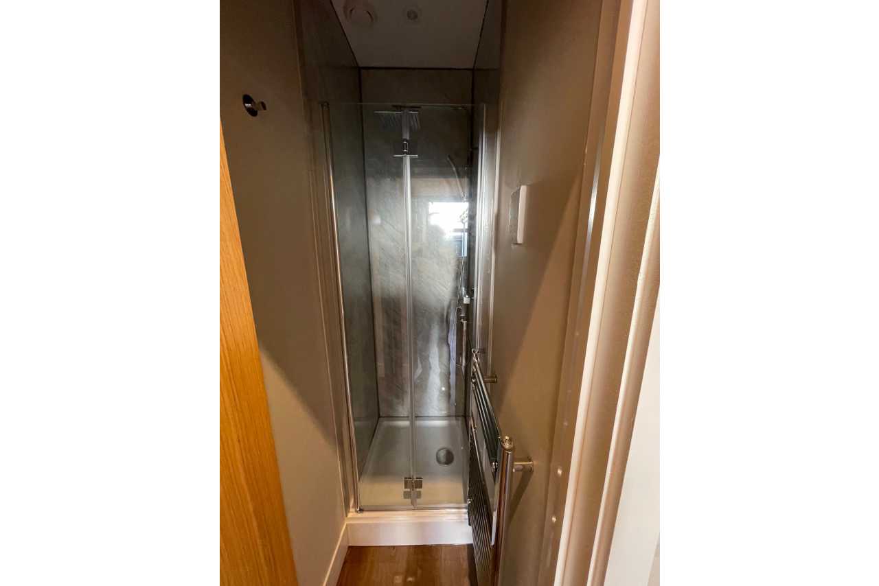 A narrow bathroom with a glass shower enclosure, wall-mounted shower fixtures and wood-effect flooring.