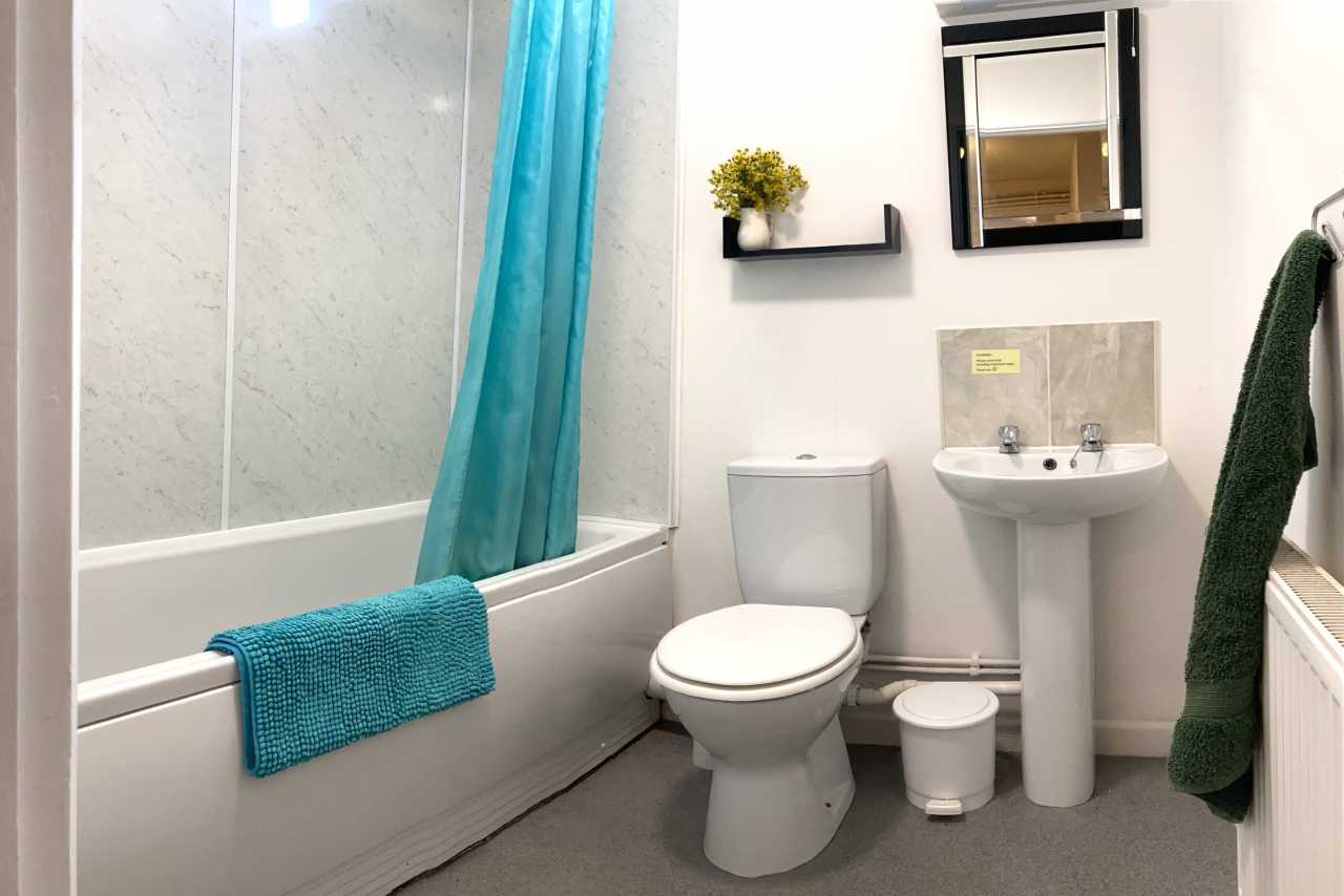 A clean bathroom with a bathtub, blue shower curtain, toilet, pedestal sink, mirror and radiator.