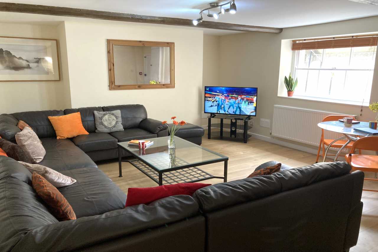 A furnished communal living room with sofas, a coffee table, television, dining table and windows.