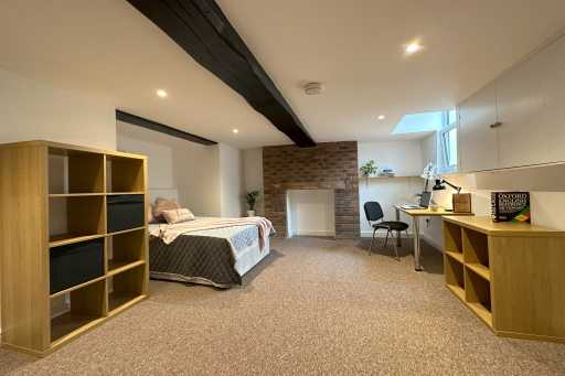 A furnished bedroom with a bed, shelving, desk, chairs, exposed brickwork and a small high window.