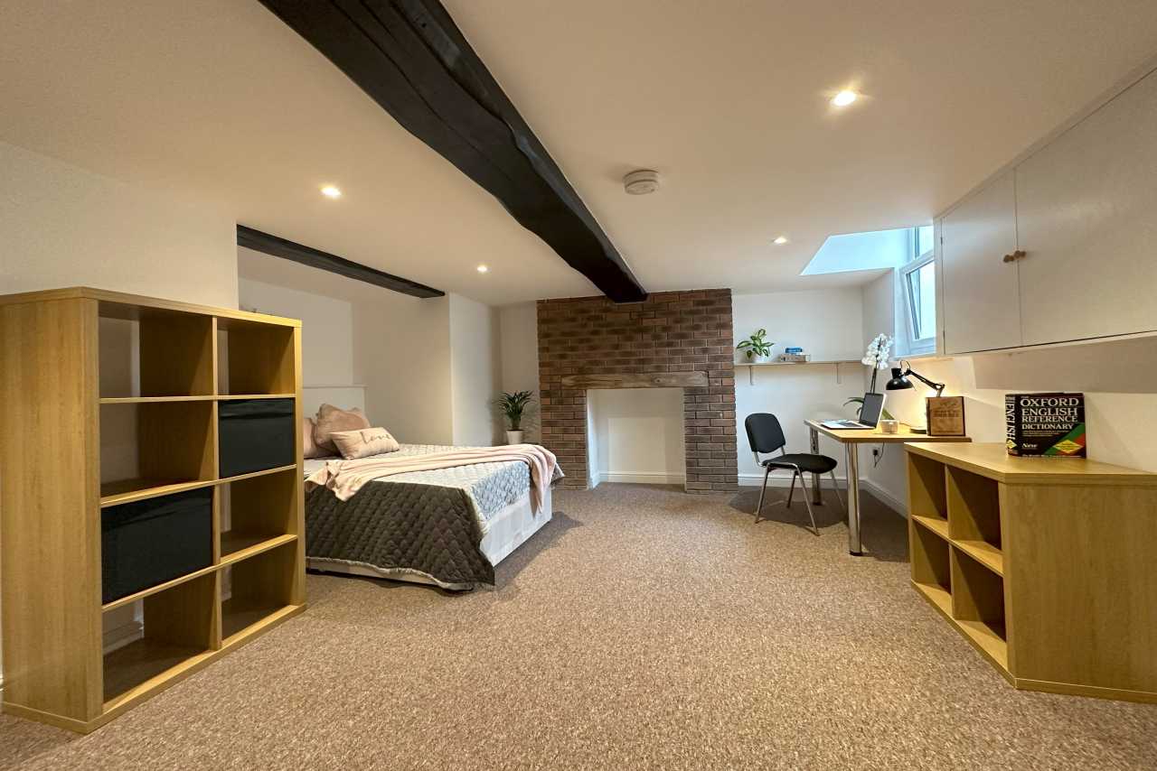 A furnished bedroom with a bed, shelving, desk, chairs, exposed brickwork and a small high window.