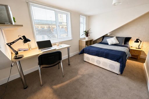 A furnished bedroom with a bed, desk, chair, lamps and windows.