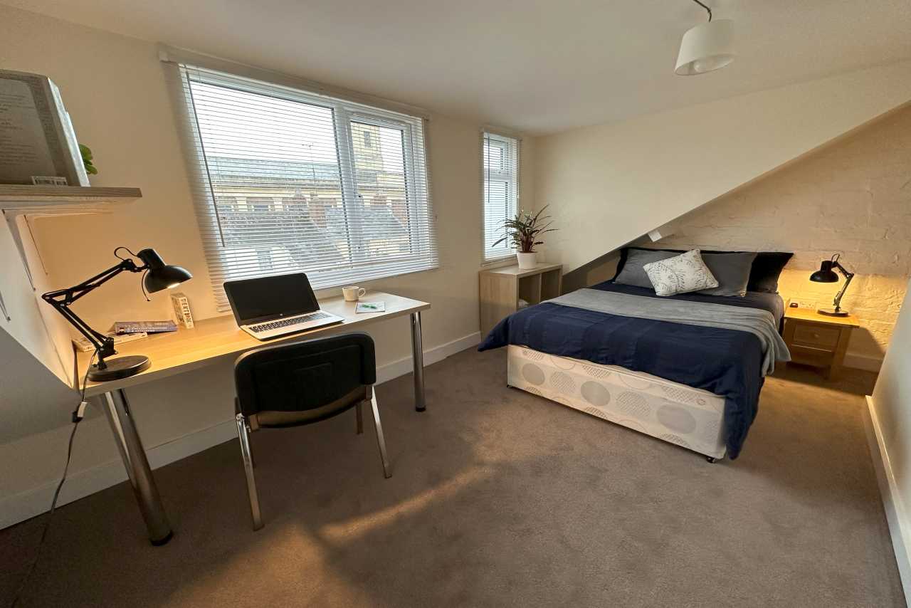 A furnished bedroom with a bed, desk, chair, lamps and windows.