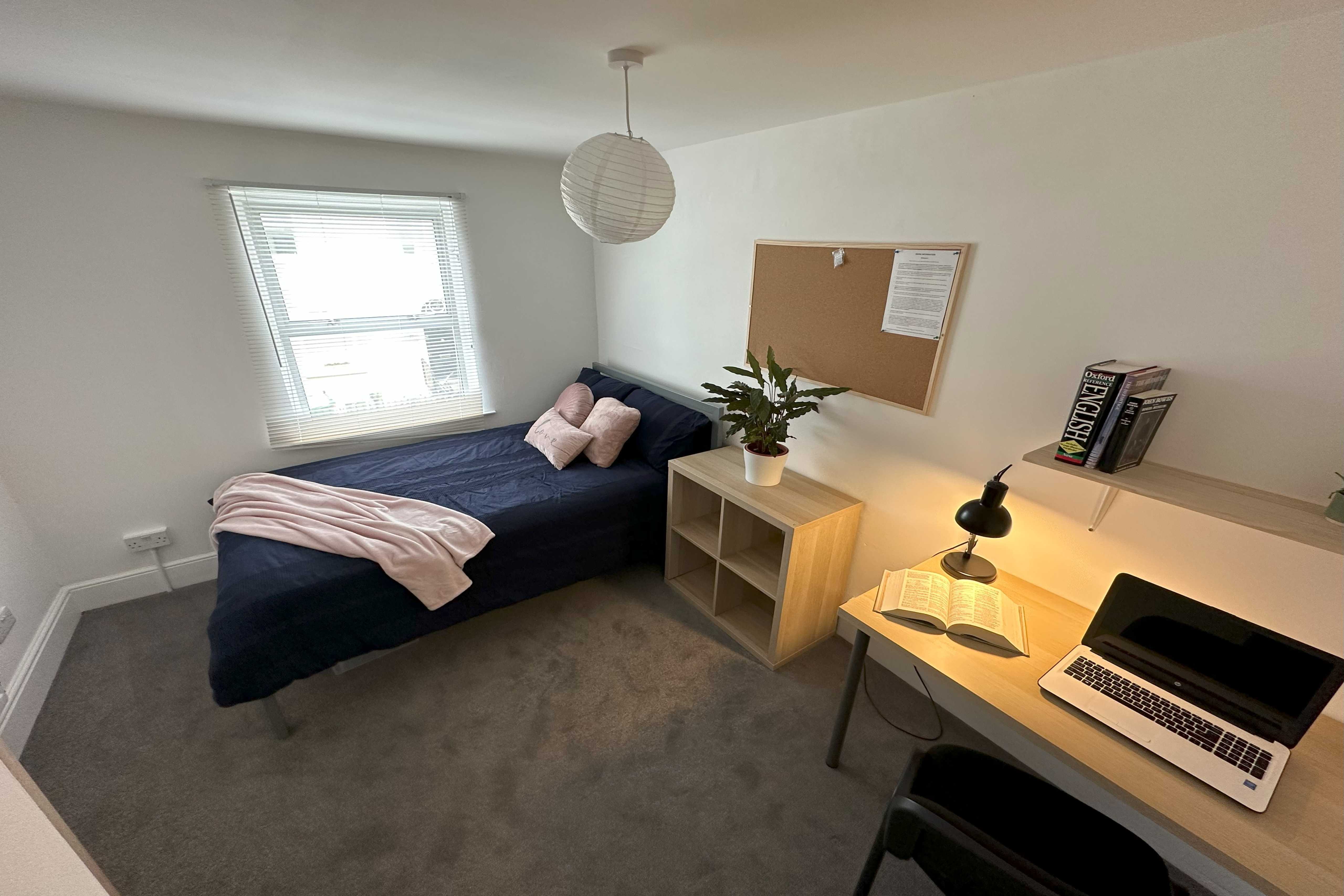 A furnished bedroom with a single bed, desk, chair, shelving, window and corkboard.