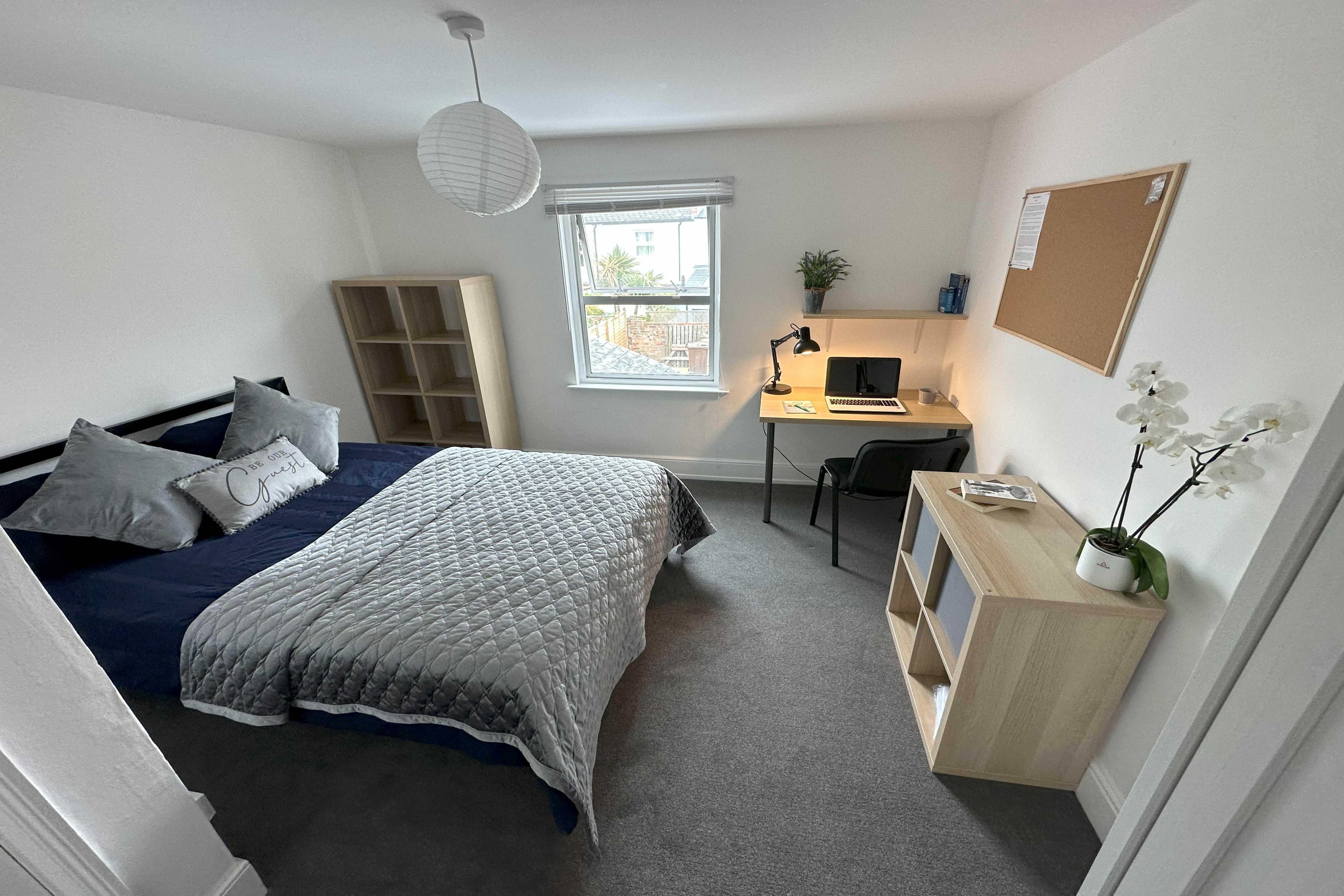 A furnished bedroom with a double bed, desk, chair, shelving, window and potted plants.
