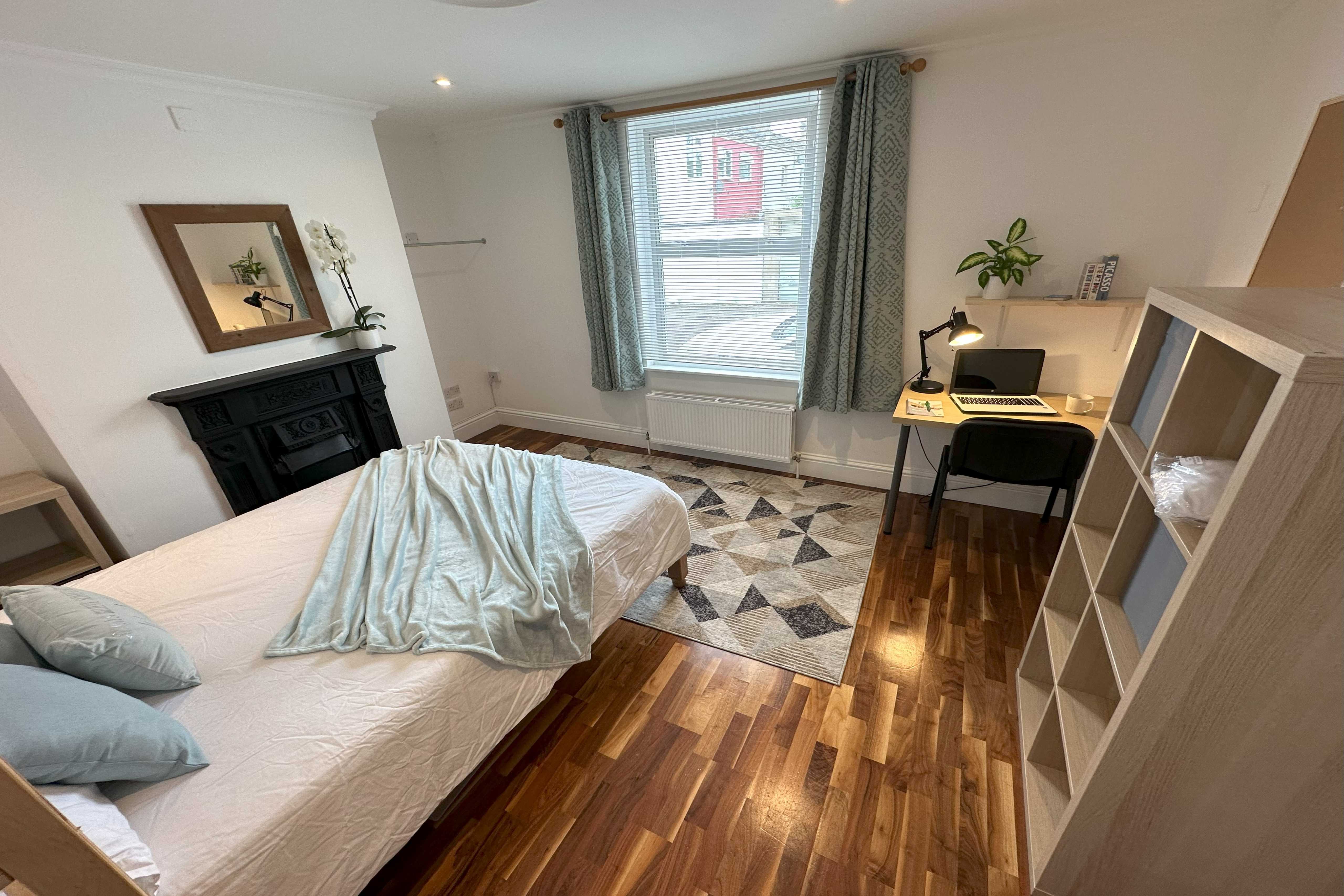 A furnished bedroom with a bed, fireplace, desk, chair, shelving and a window with curtains.