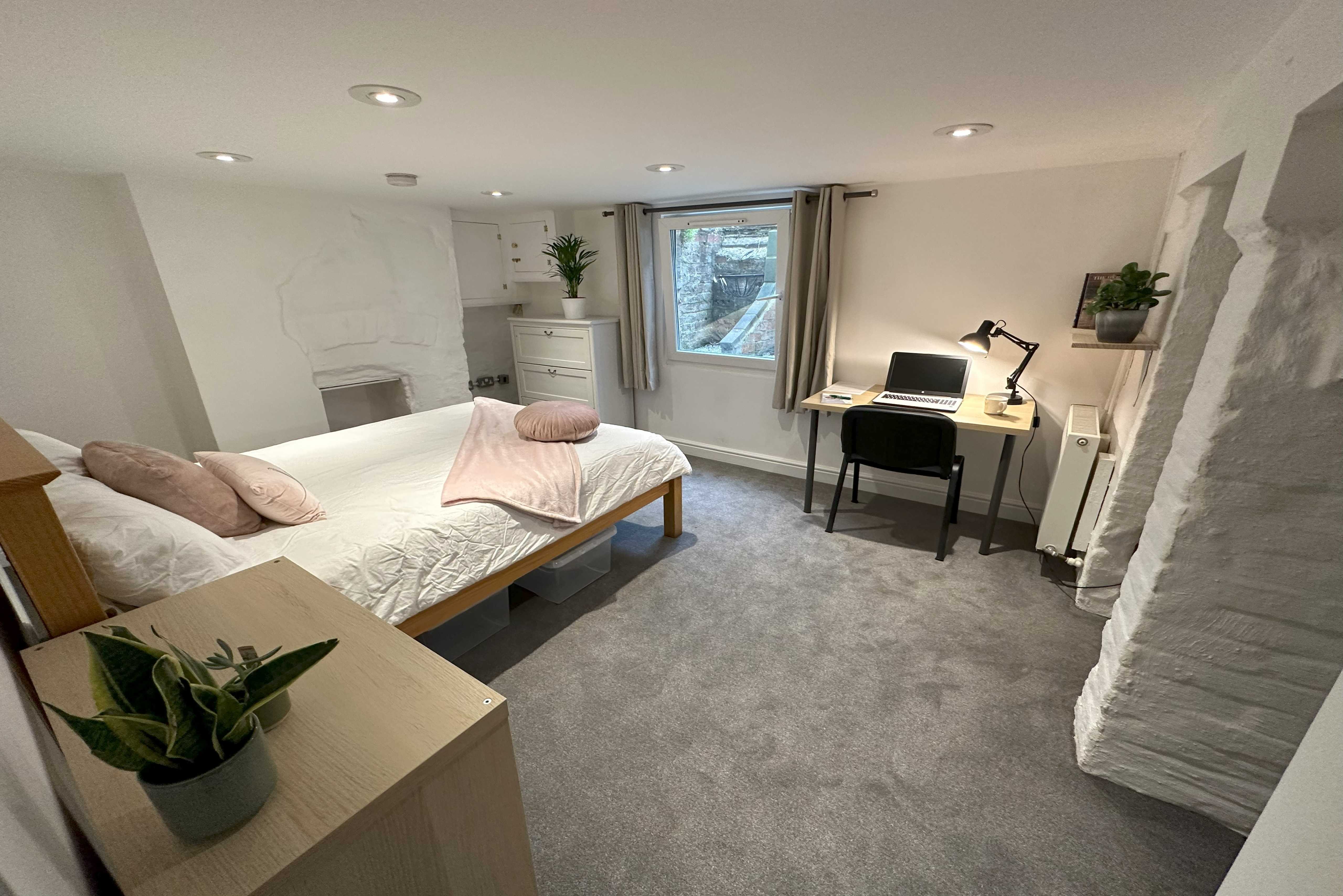 A furnished bedroom with a bed, desk and chair, window, shelving, and carpeted floor.