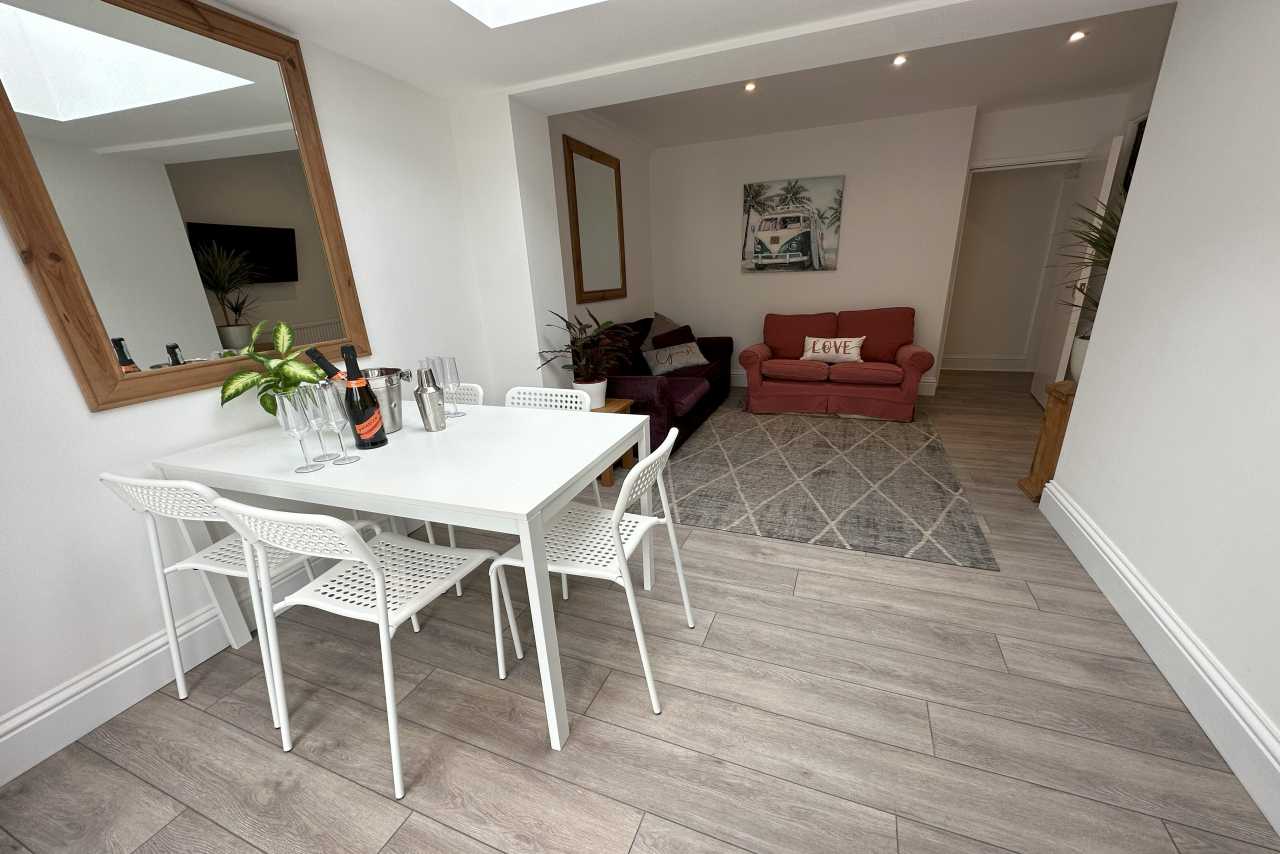 A bright furnished dining and sitting area with a white table and chairs, sofas, wall art, a large mirror and wood-effect flooring.