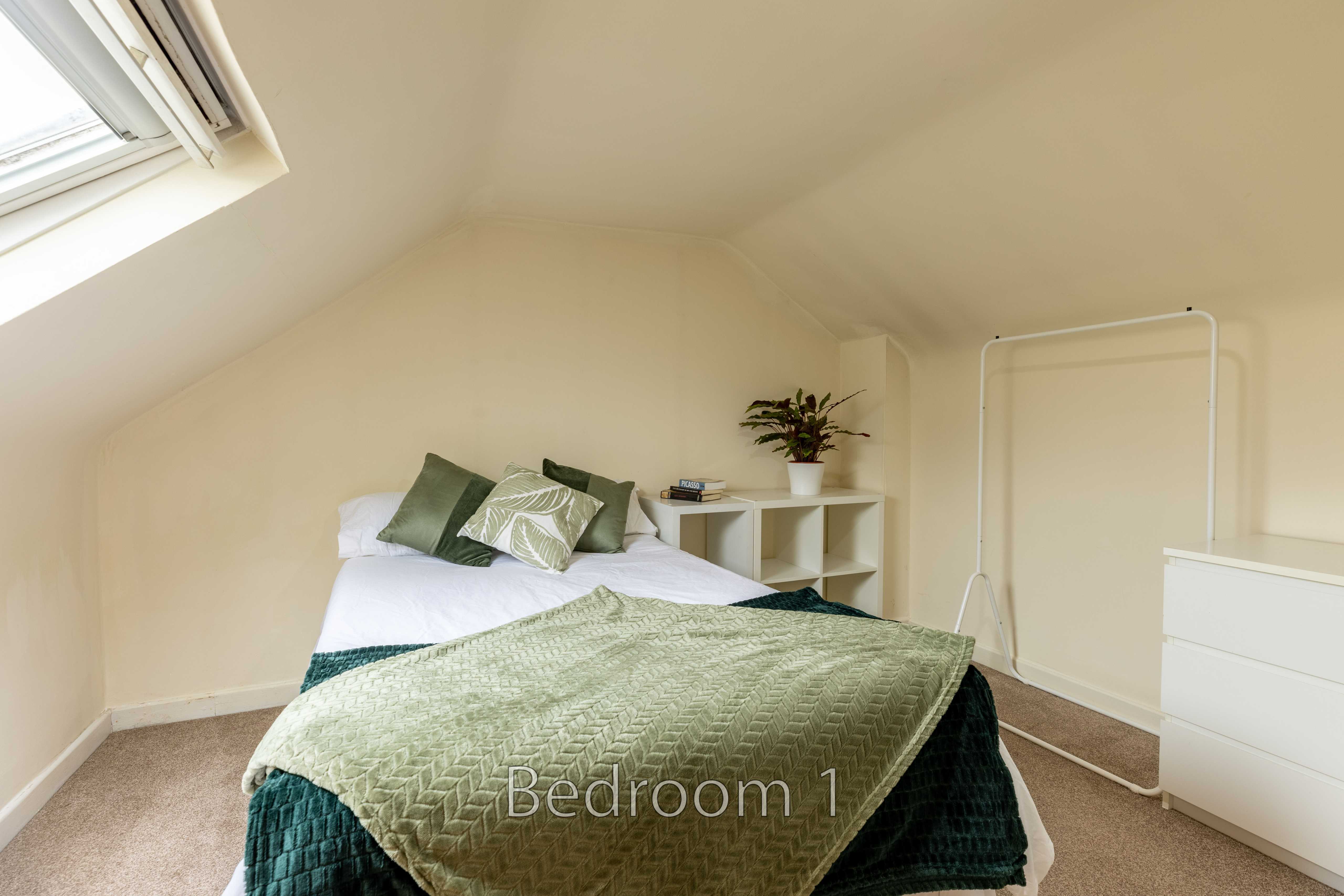 A furnished attic bedroom with a double bed, green bedding, bedside shelving, a chest of drawers and a skylight.