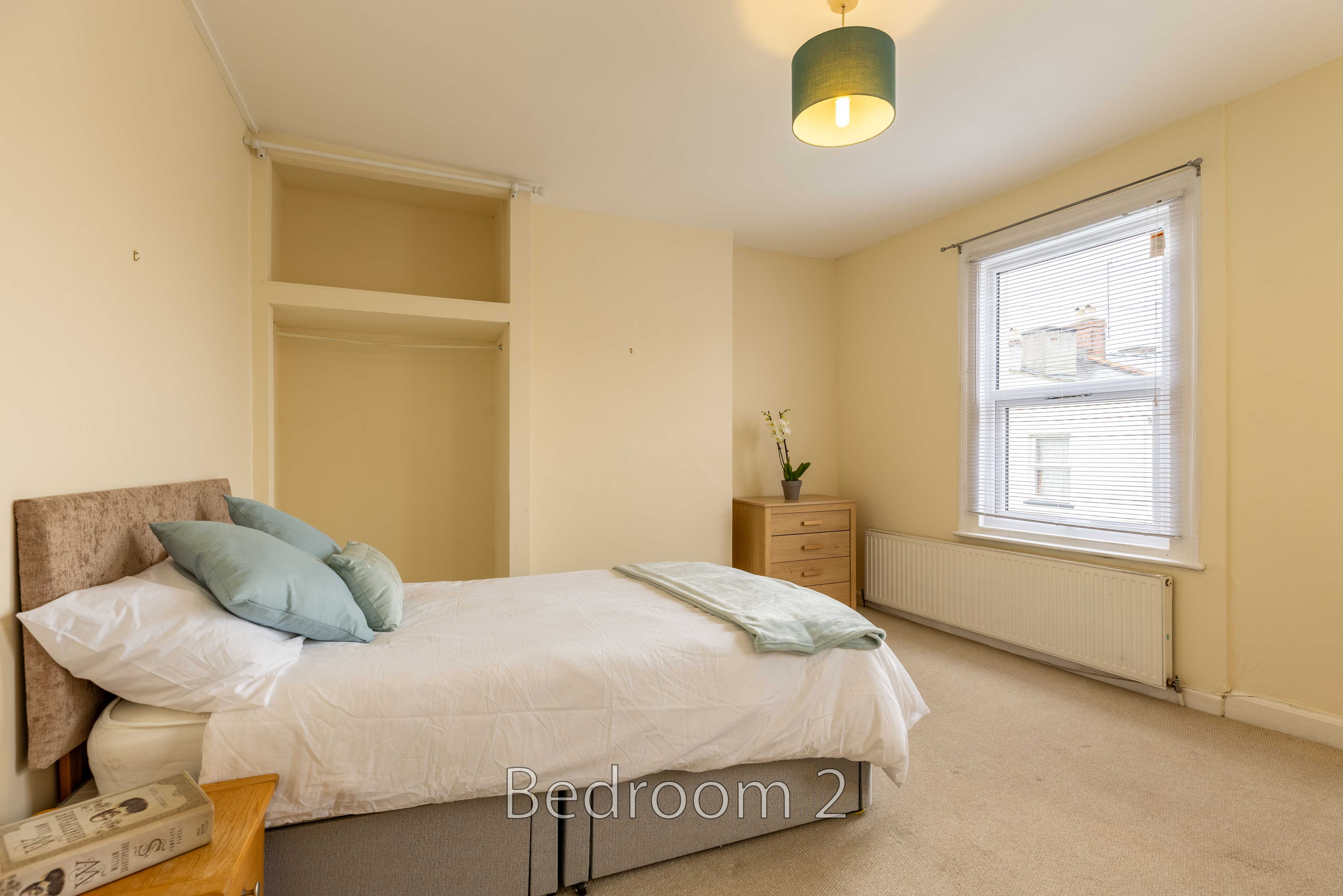 A furnished bedroom with a double bed, built-in shelving, chest of drawers, window and radiator.