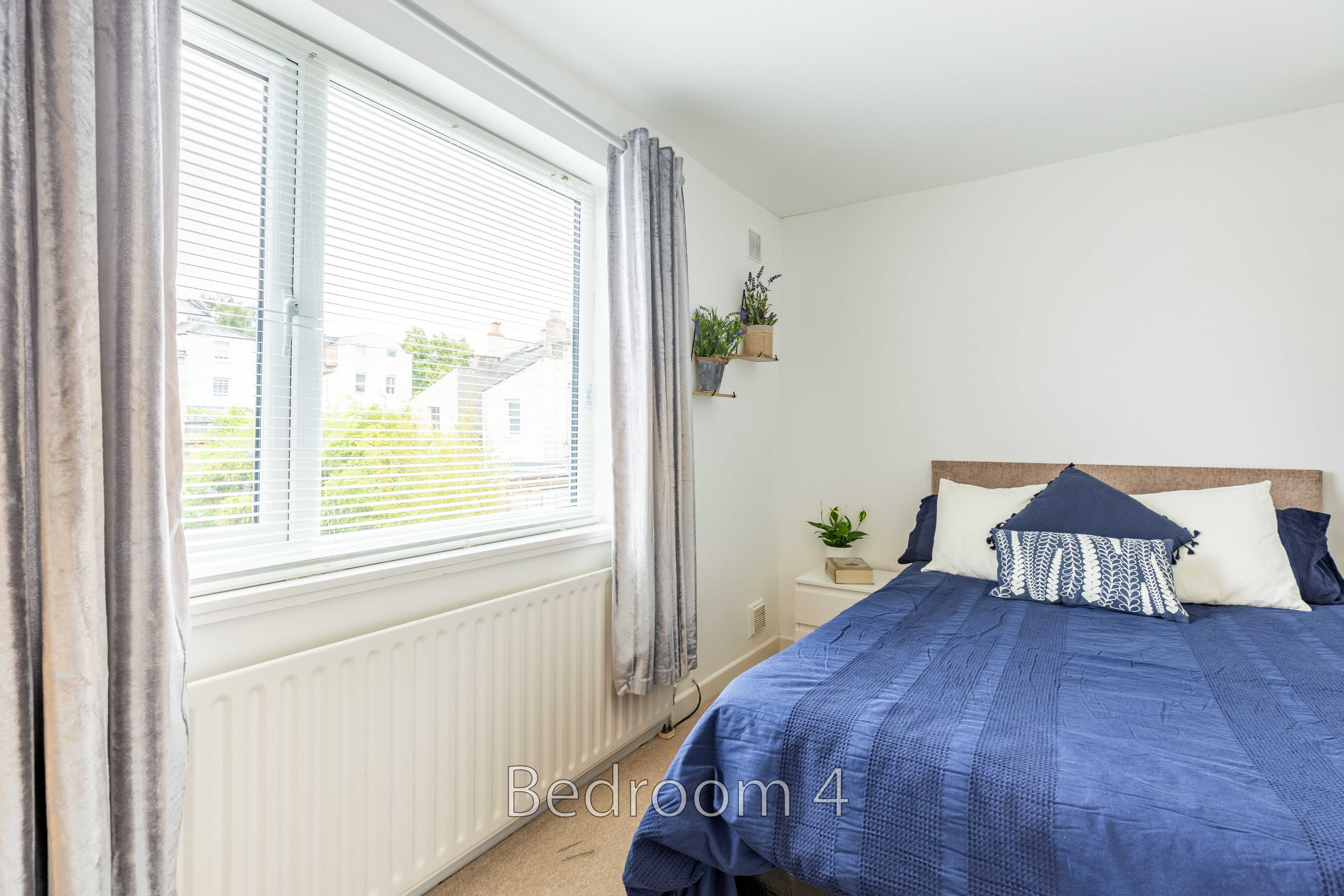 A bedroom with a double bed covered in blue bedding, a large window, curtains, radiator and small wall shelves.