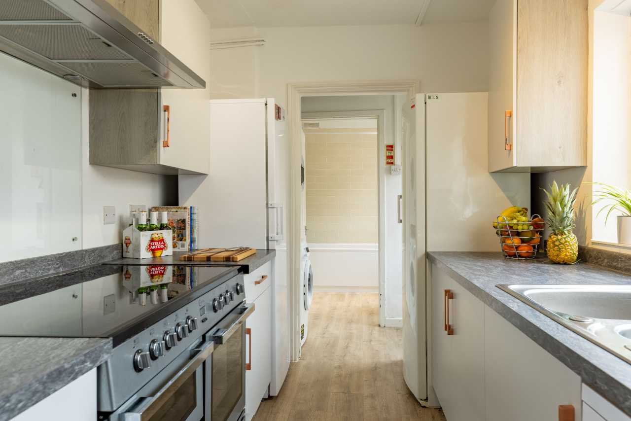 A bright kitchen with white cabinets, a large cooker, countertops, a sink, and a doorway leading to a tiled room.