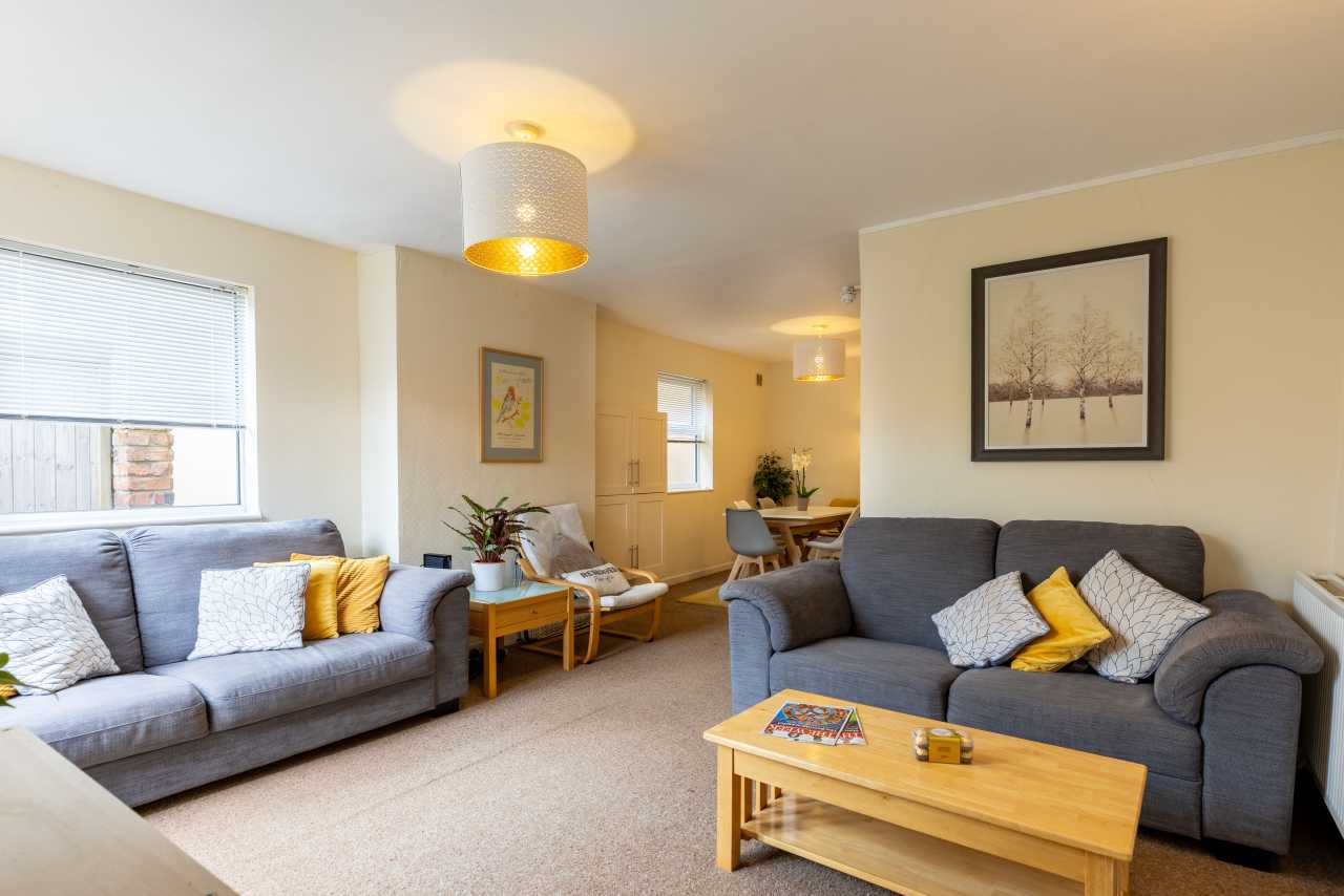Bright living room with two grey sofas, a wooden coffee table, armchairs, framed artwork, and windows with blinds.