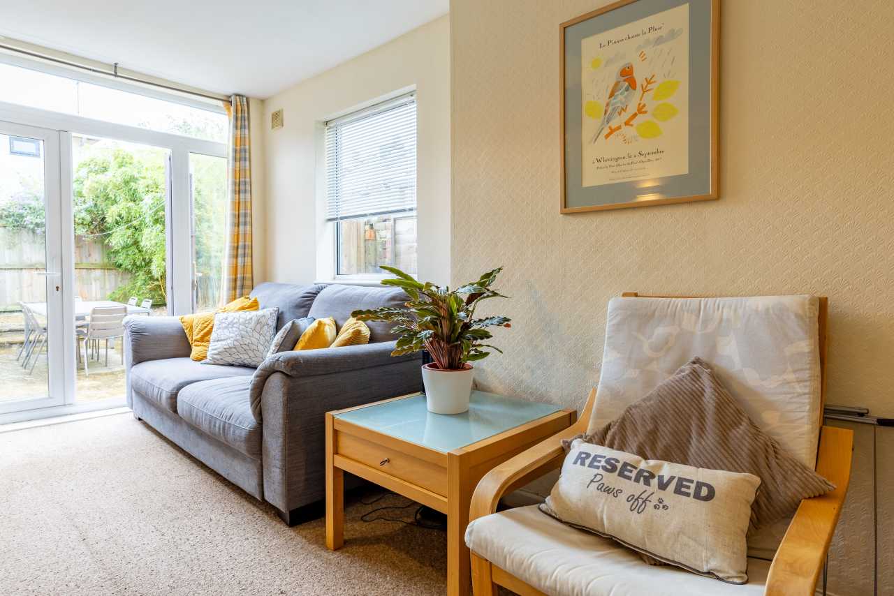 Bright furnished sitting area with a grey sofa, armchair, coffee table, framed artwork and glass doors opening to a garden.
