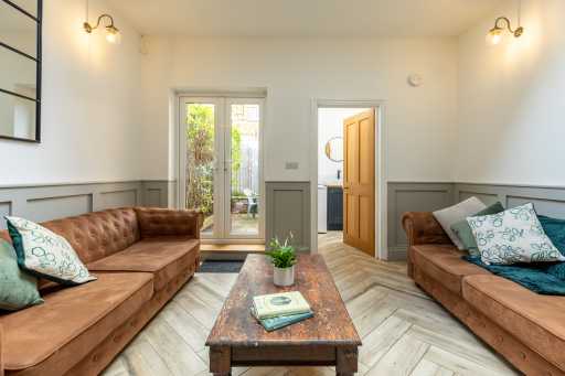 Bright living room with two brown sofas, a central wooden coffee table, wall panelling, and glass doors opening to greenery.