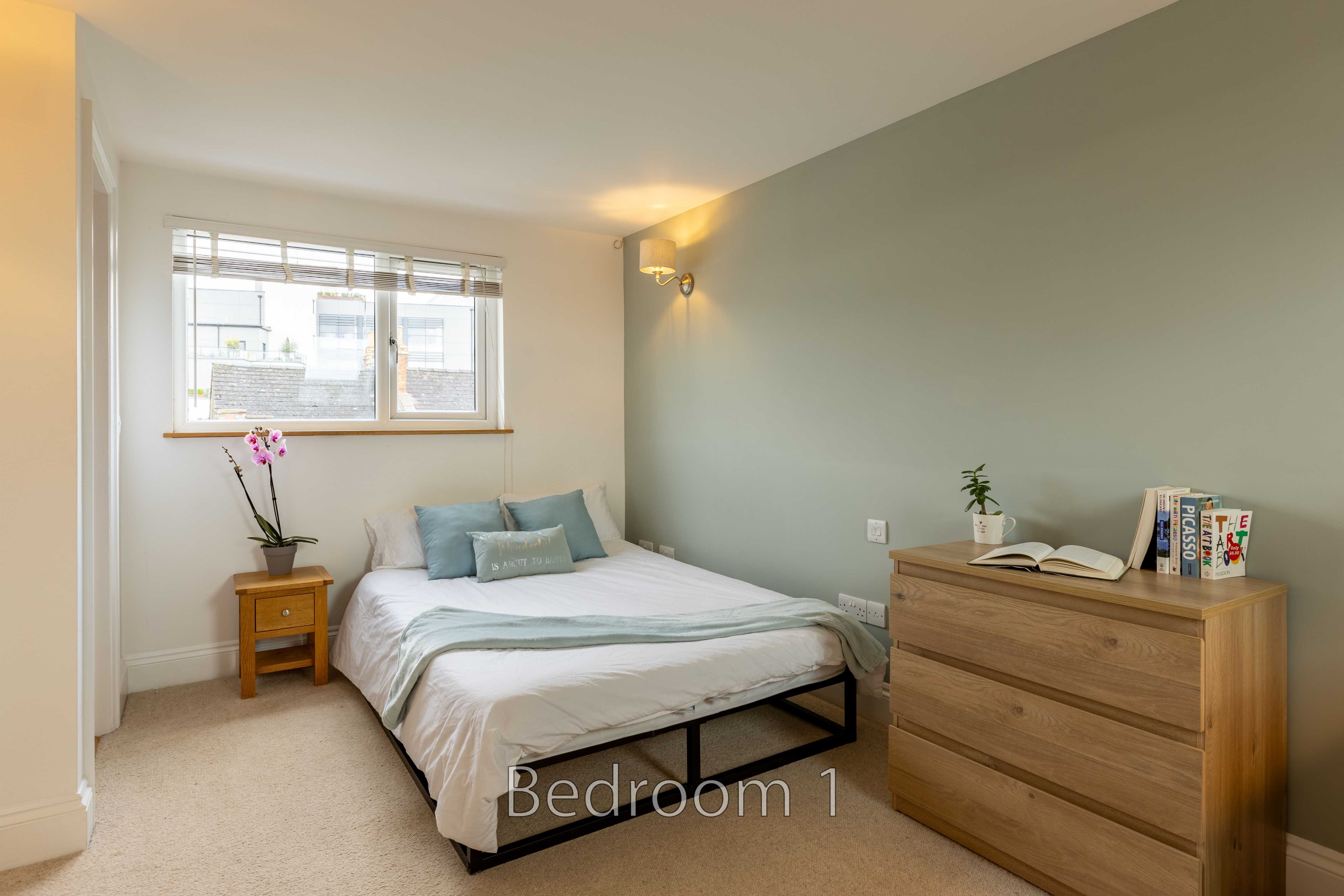 A furnished bedroom with a double bed, bedside table, chest of drawers, window and wall light.