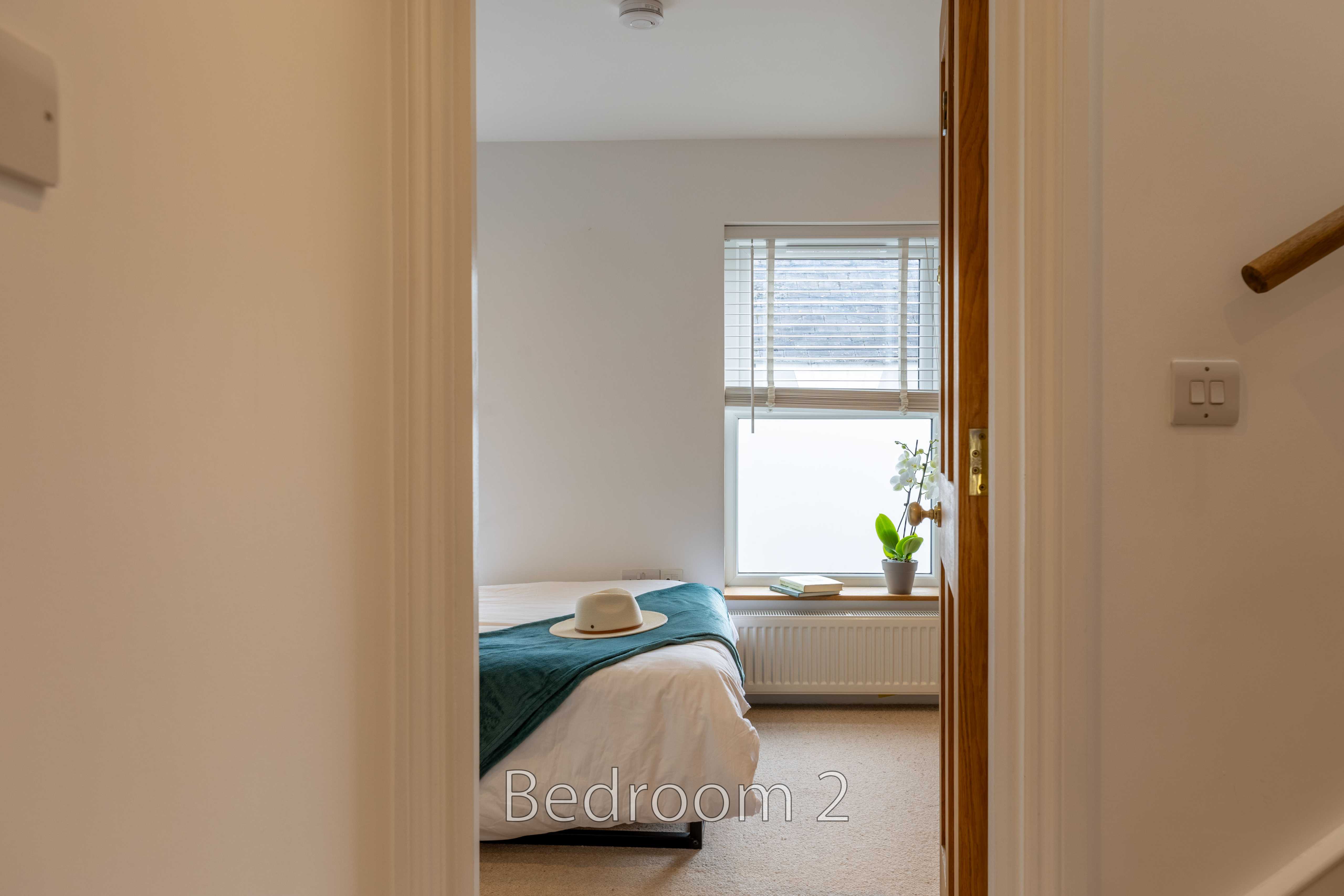 A bedroom viewed through an open doorway, with a bed, window, radiator and potted plant.