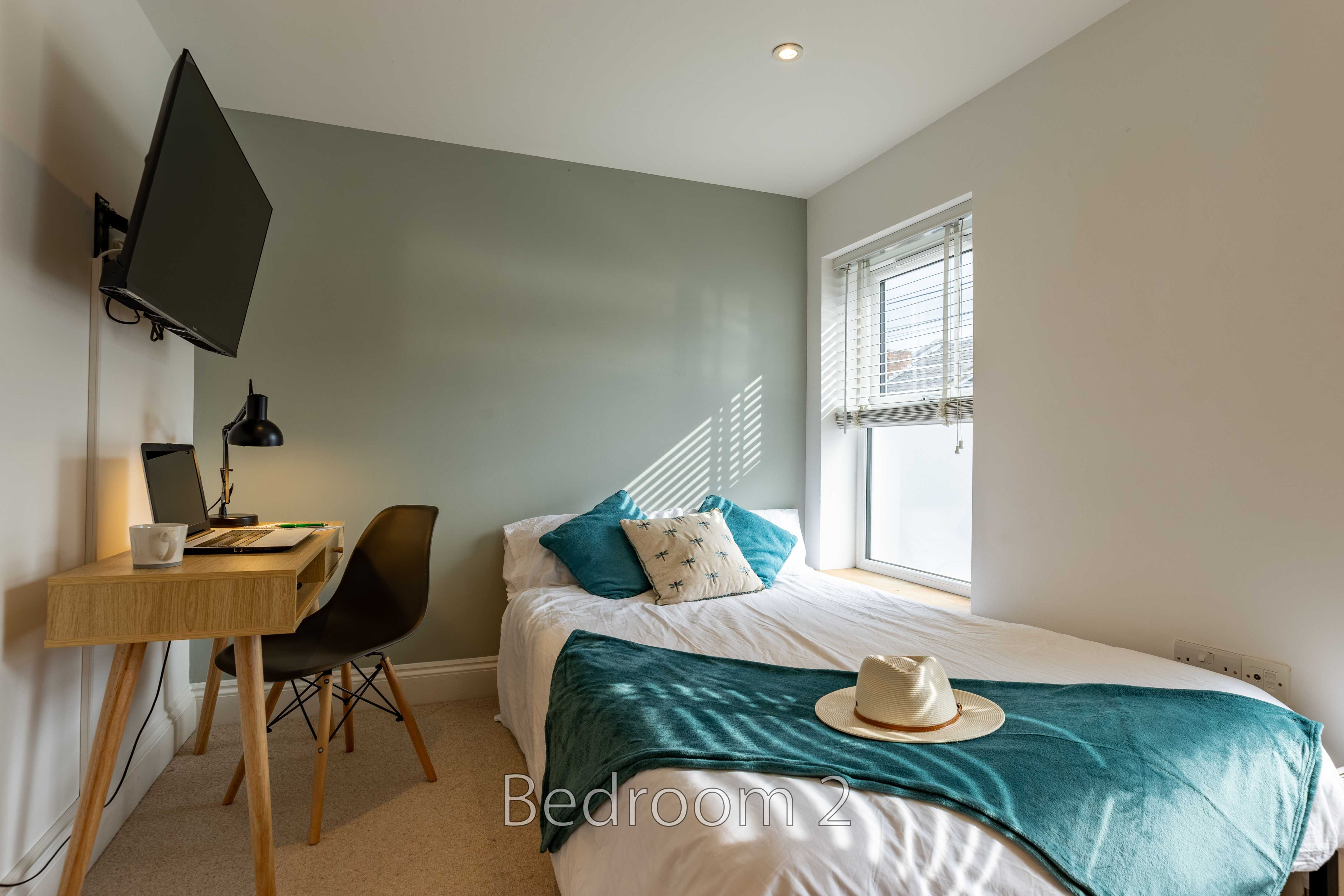A furnished bedroom with a single bed, desk, chair, wall-mounted television and window.