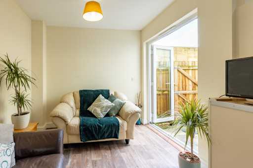 Bright sitting area with an armchair, sofa, television, potted plants and an open glass door to a fenced outdoor area.