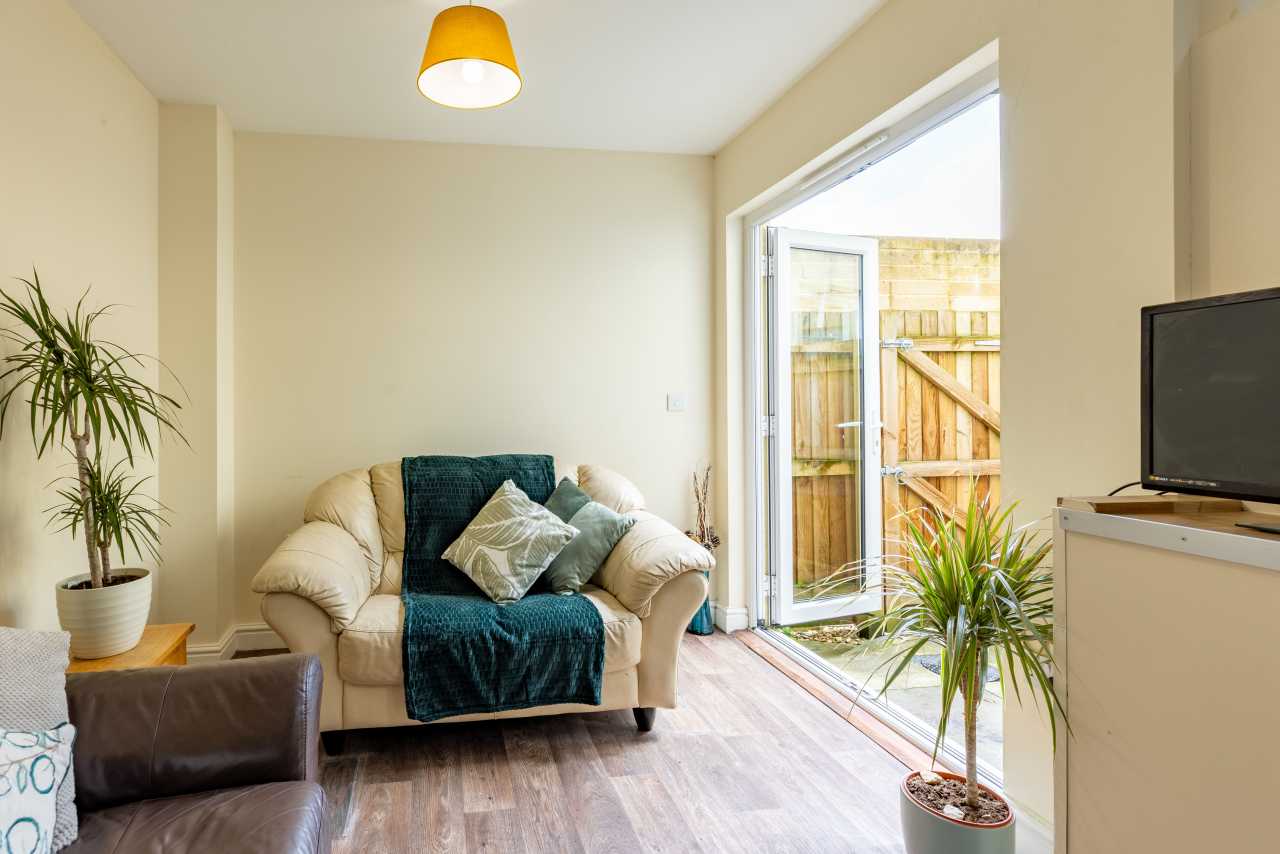 Bright sitting area with an armchair, sofa, television, potted plants and an open glass door to a fenced outdoor area.