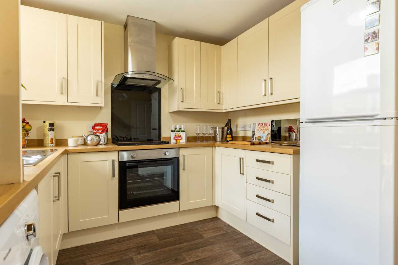 A fitted kitchen with cream cabinets, an oven and hob, a range hood, countertops, and a tall white refrigerator.