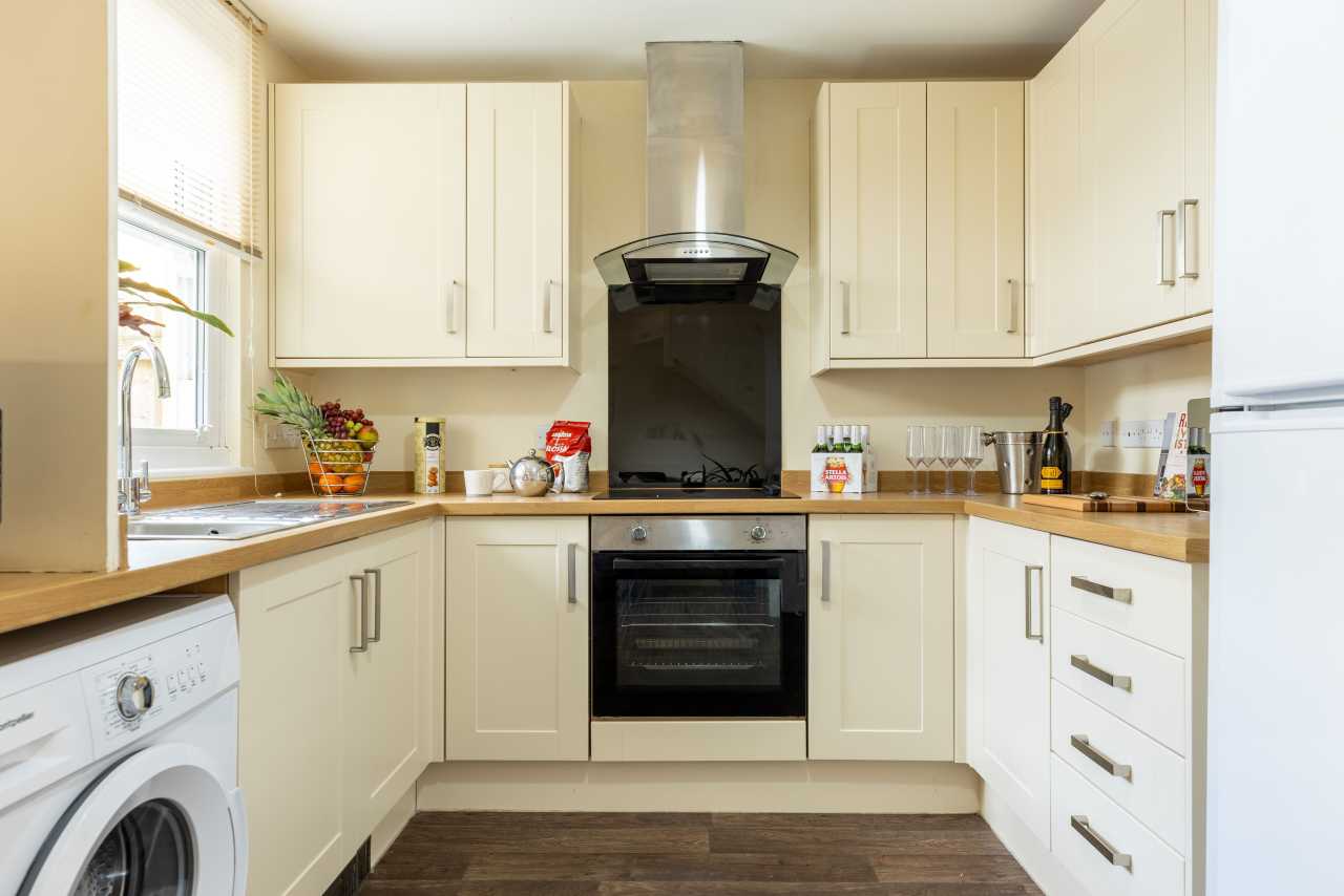 Bright fitted kitchen with cream cabinets, an oven, extractor hood, sink, washing machine and countertop appliances.