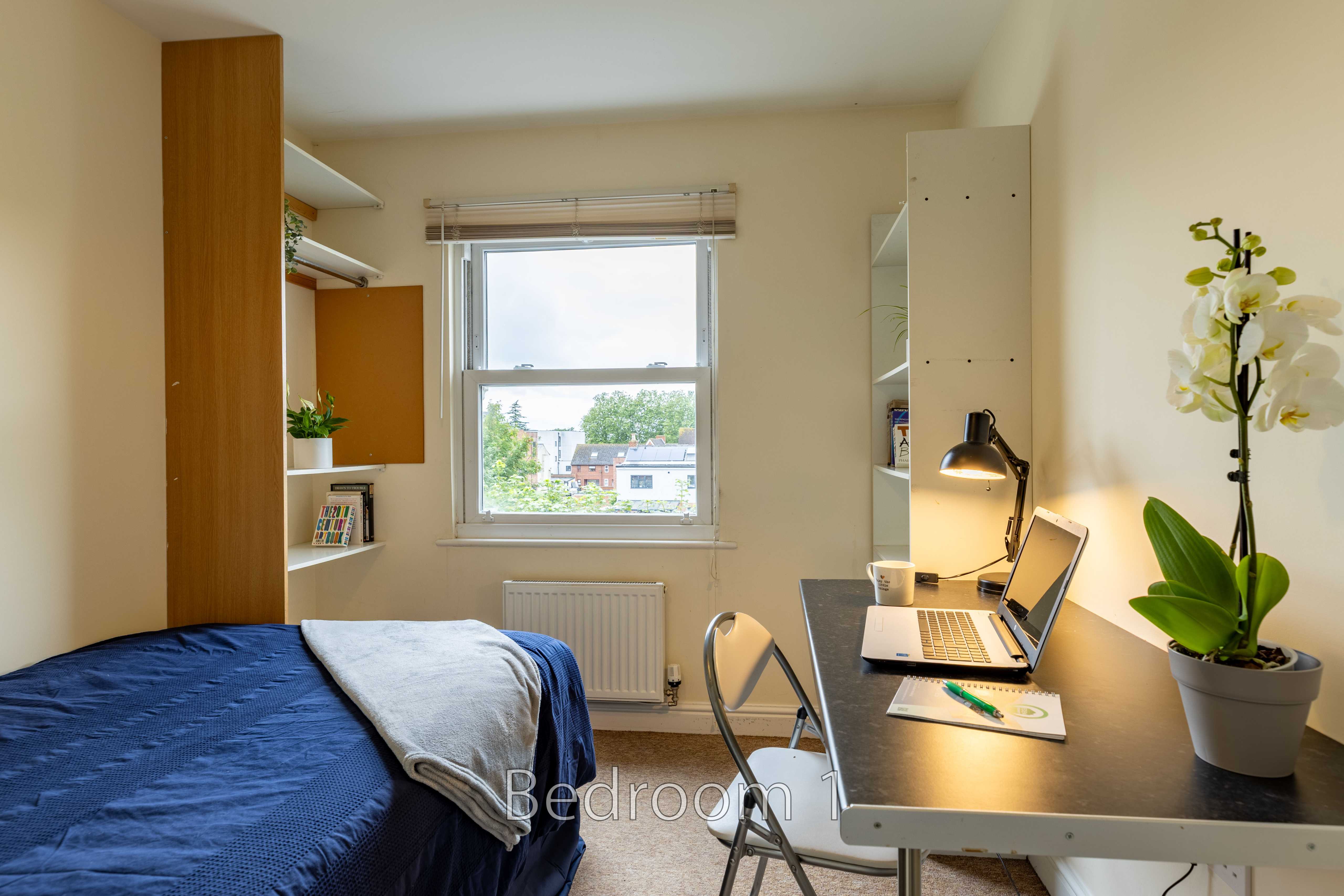 A furnished bedroom with a bed, desk, chair, window, shelving and a potted orchid.
