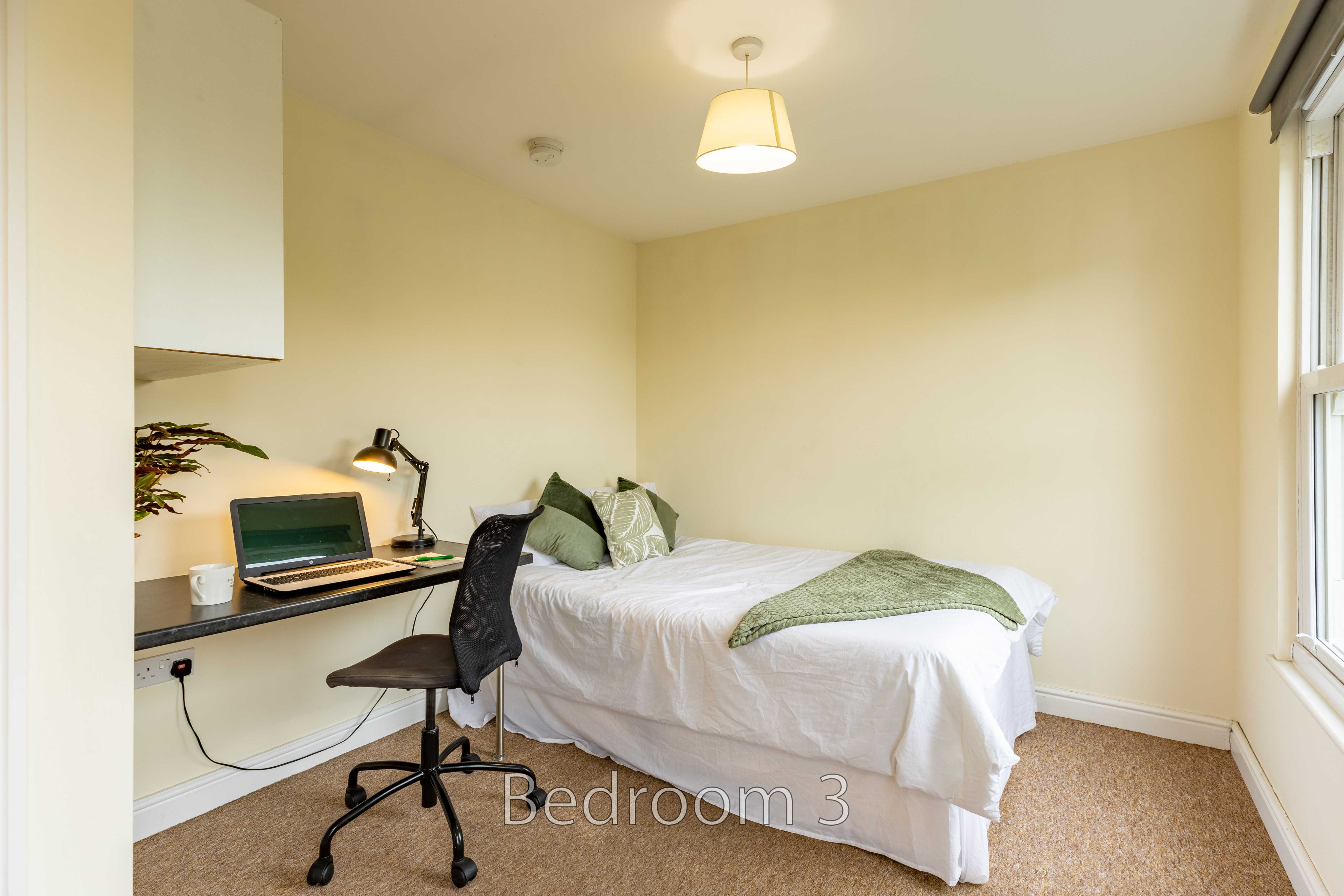 A bedroom with a single bed, desk, chair, computer and window.