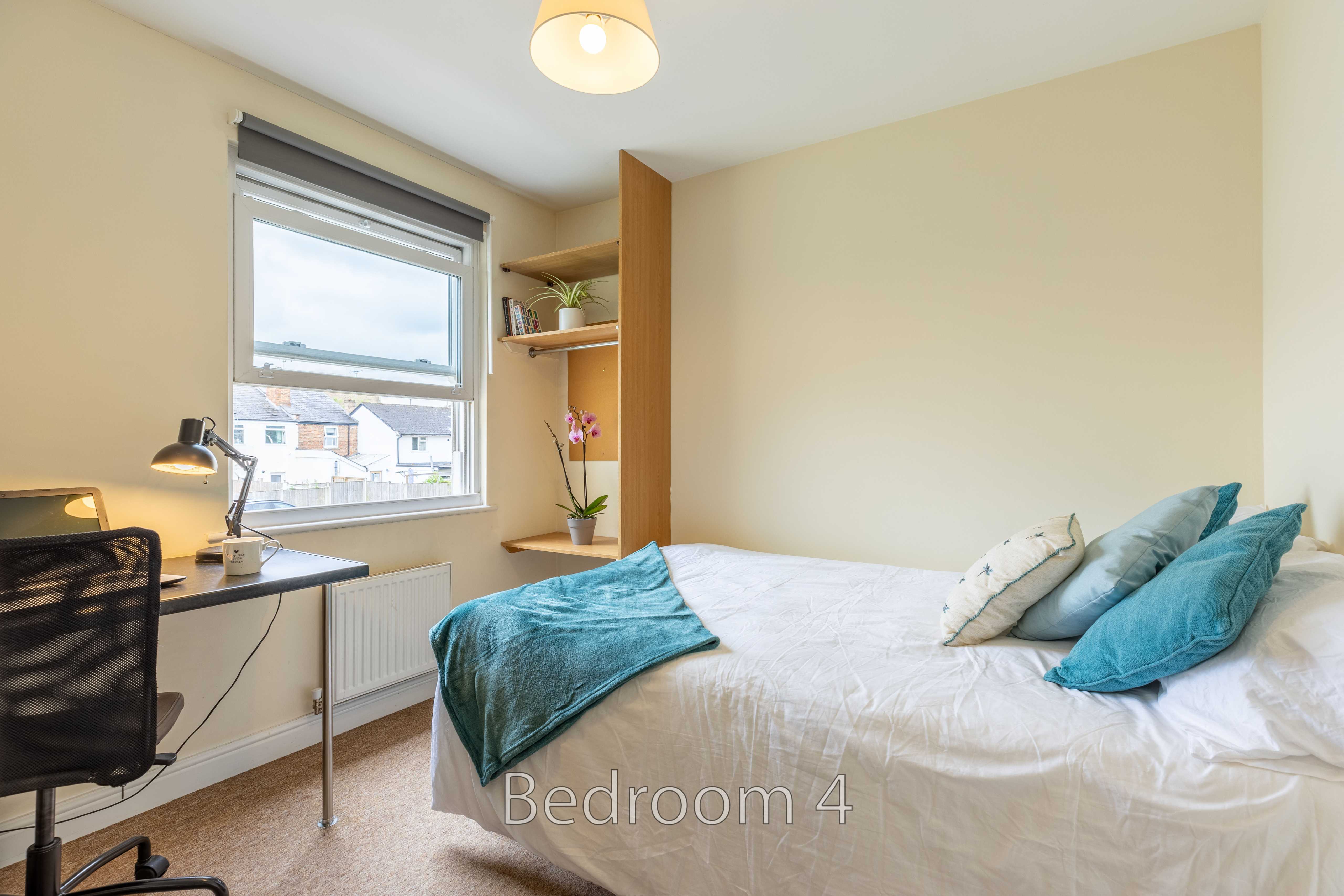 A furnished bedroom with a double bed, blue cushions, a desk, shelving and a window.