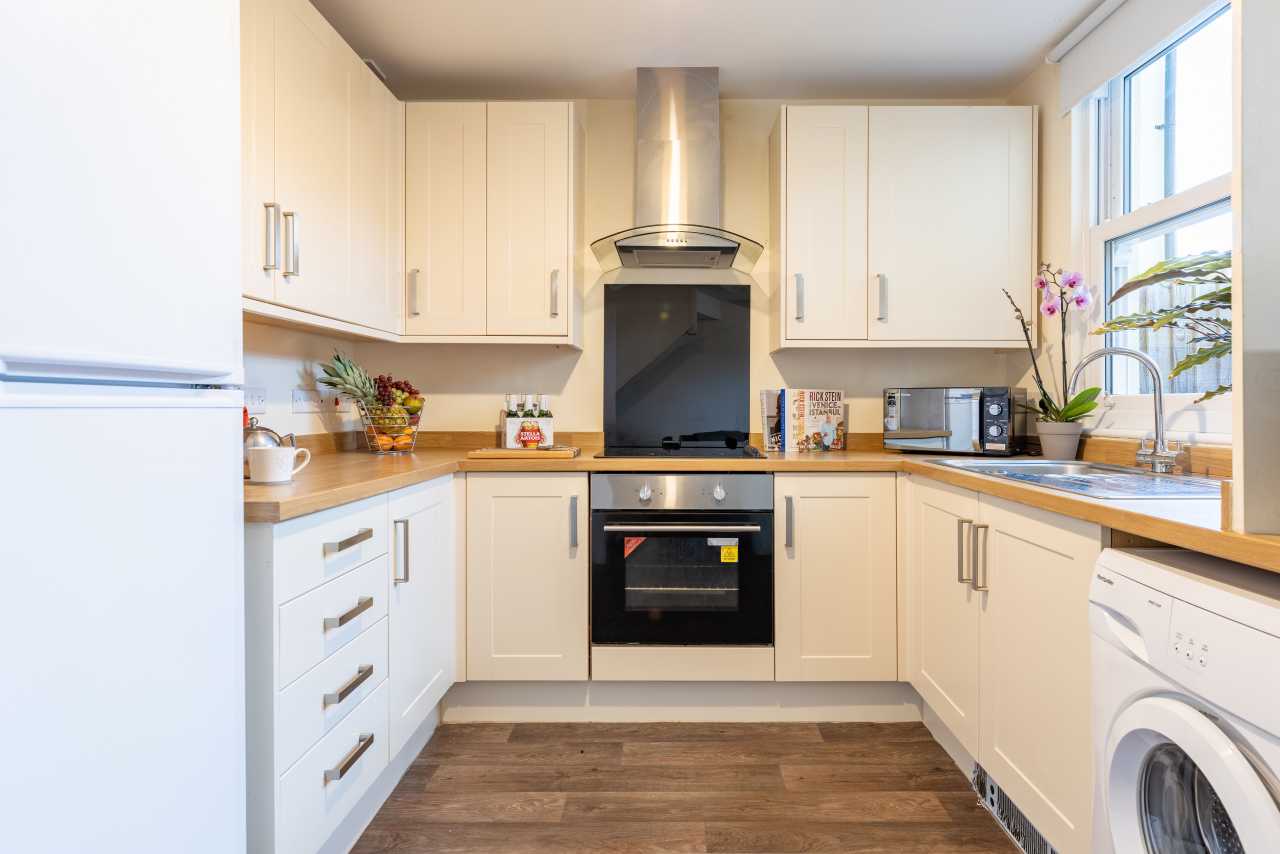 Bright fitted kitchen with cream cabinets, wood-effect flooring, cooker, sink, appliances and a large window.
