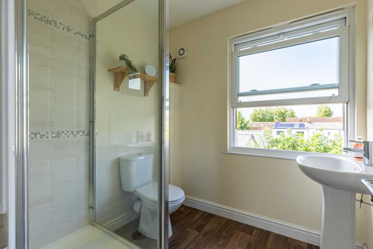 Bright bathroom with a glass shower enclosure, toilet, pedestal sink, window, tiled walls and wood-effect flooring.