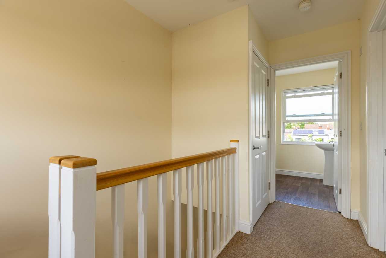 Landing area with a wooden stair railing, carpeted floor, open doors, and a bright room visible at the end.