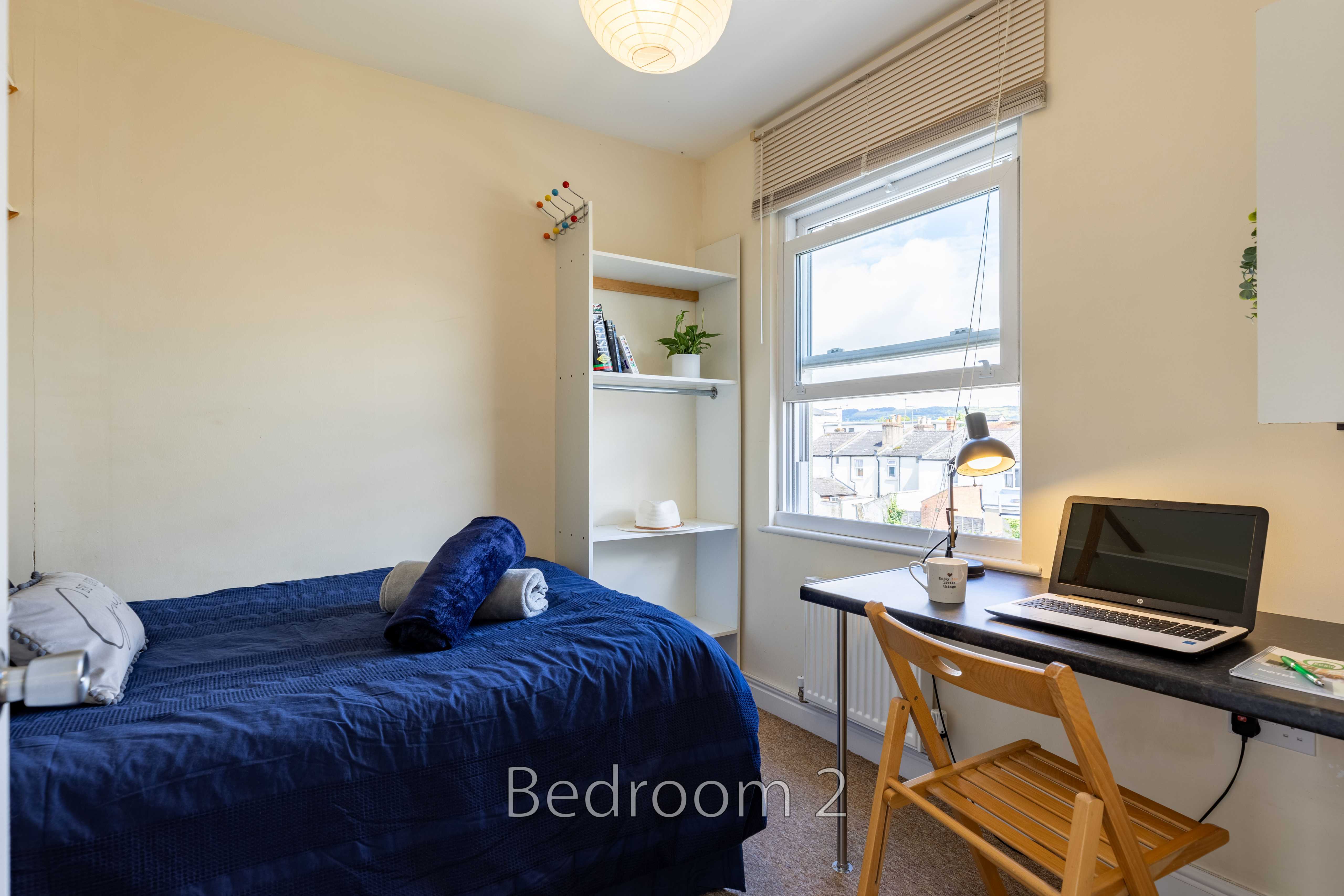 A bedroom with a blue bed, shelving, a desk, chair and window.