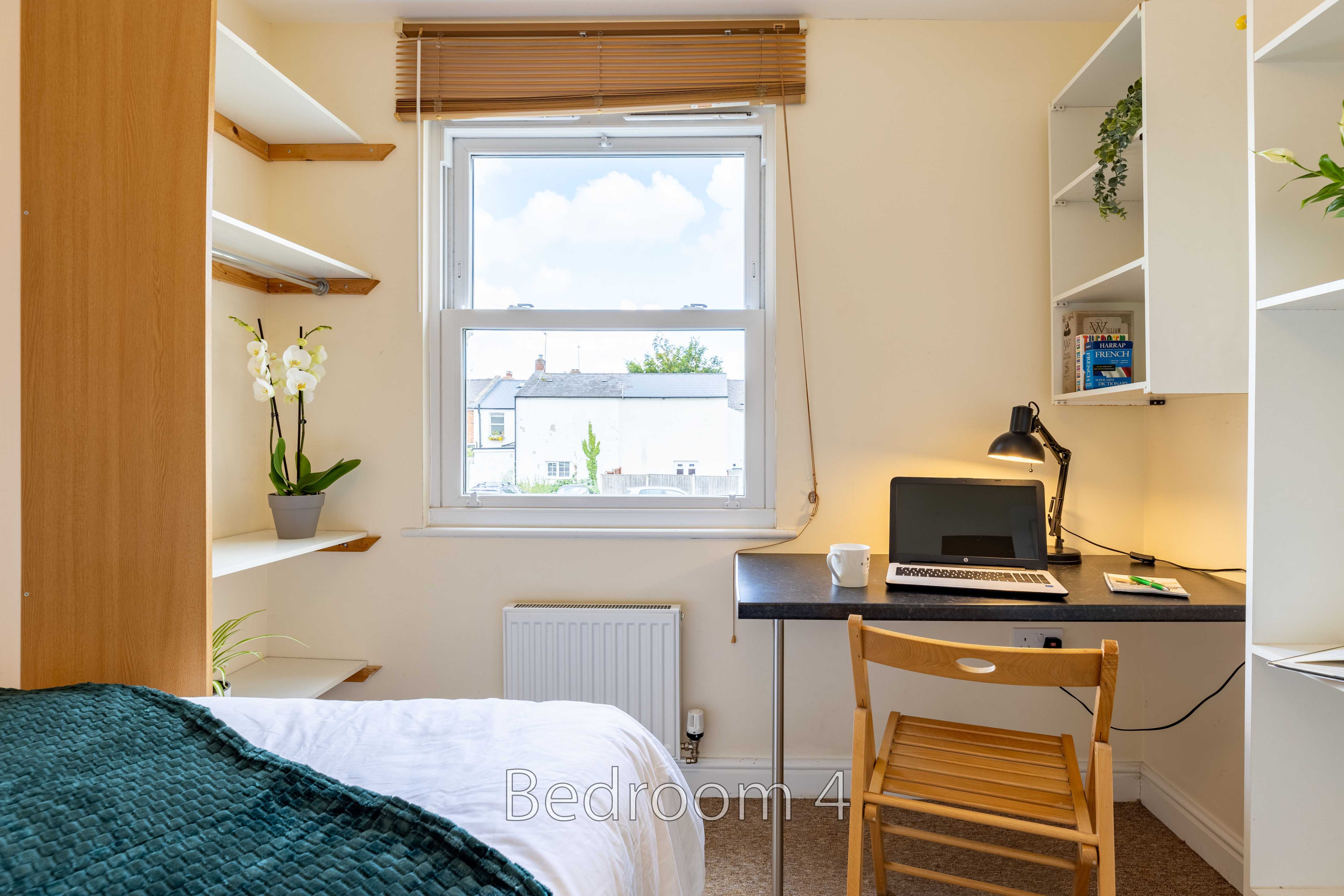 A bedroom with a bed, desk, chair, shelving, window and radiator.