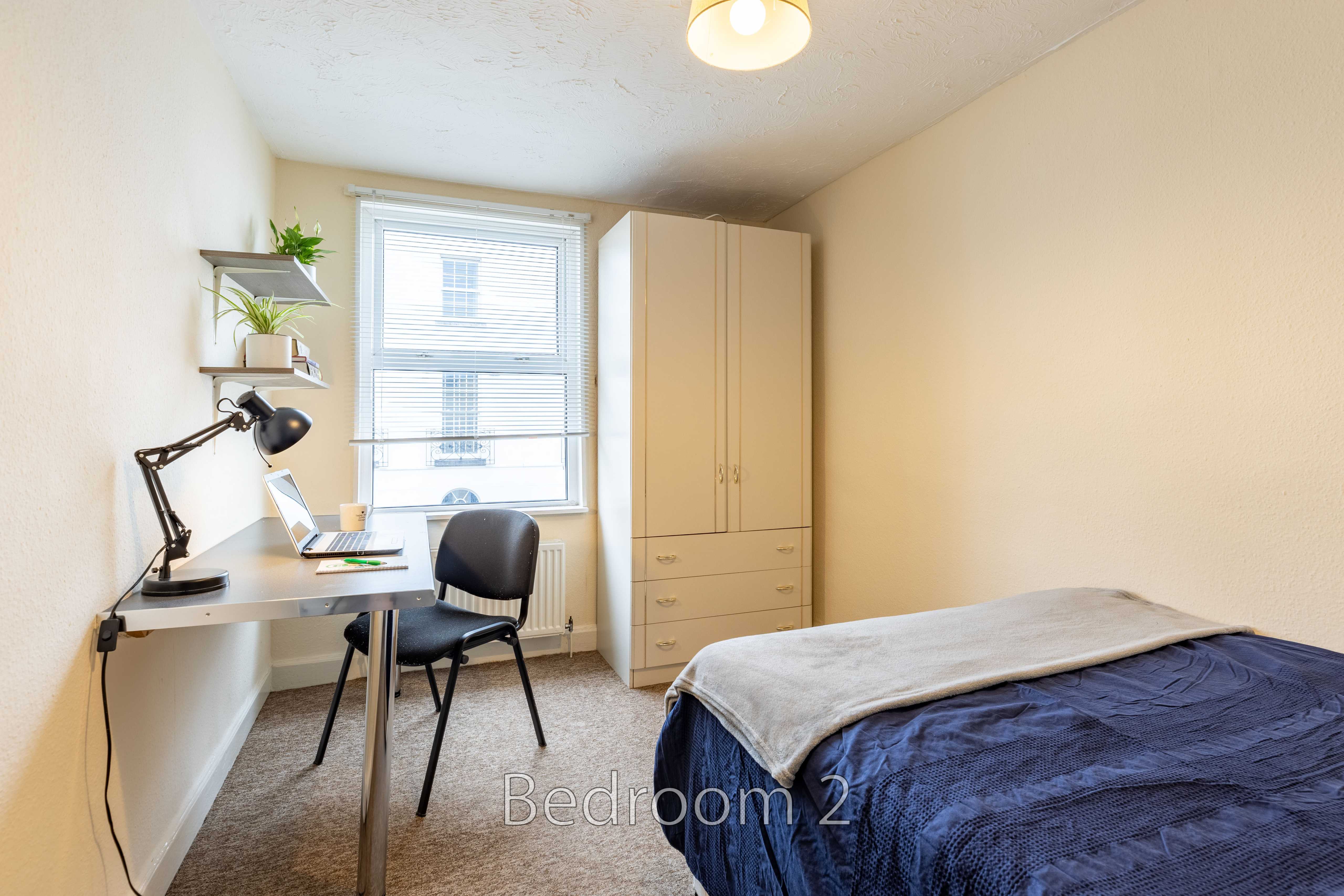 A furnished bedroom with a bed, desk, chair, wardrobe, window and shelving.