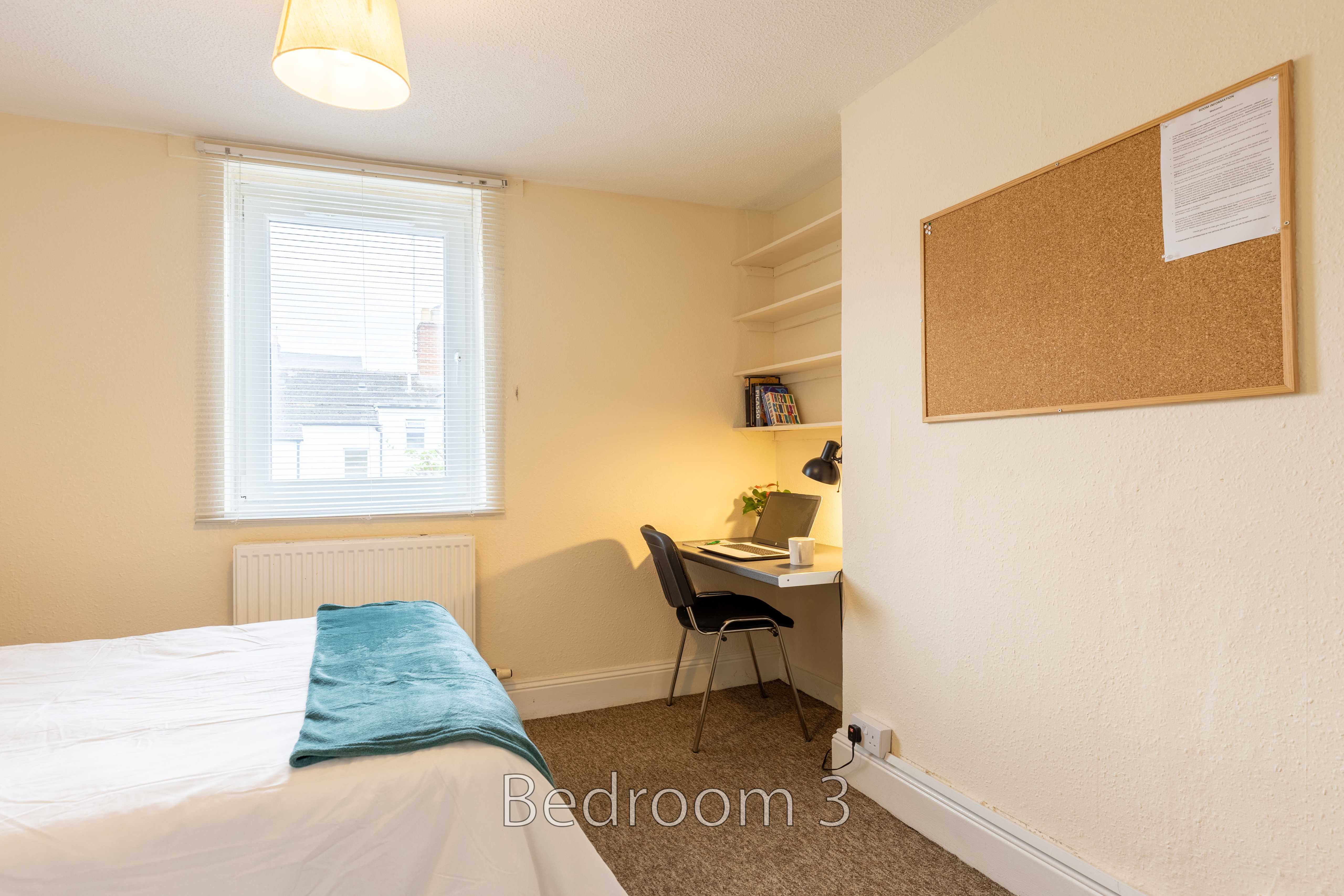 A bedroom with a single bed, desk, chair, window, shelving and a corkboard.
