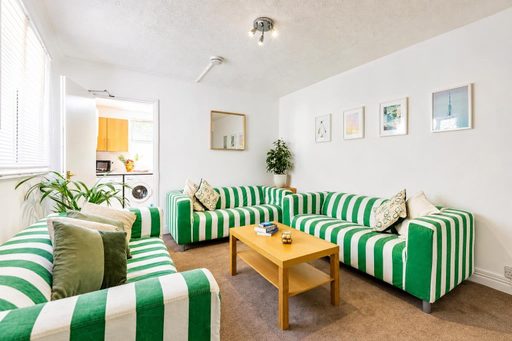 Bright living room with green-and-white striped sofas, a wooden coffee table, framed wall art, plants and a washing machine visible in the adjacent room.