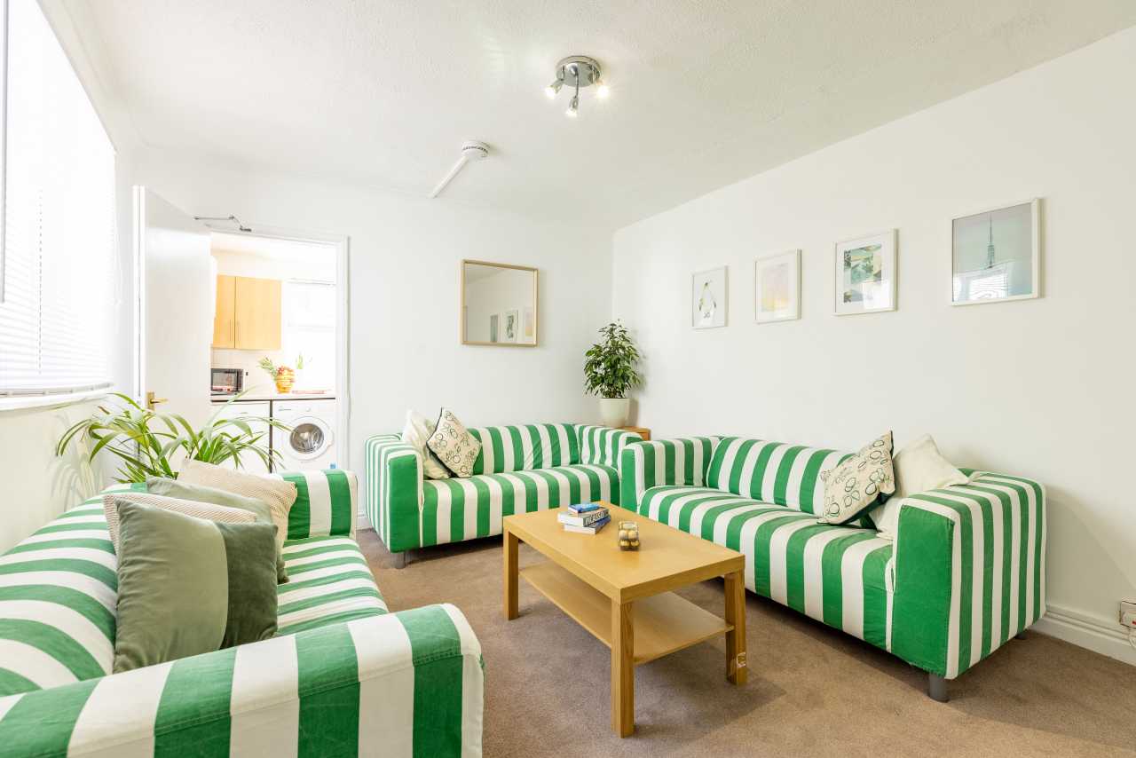 Bright living room with green-and-white striped sofas, a wooden coffee table, framed wall art, plants and a washing machine visible in the adjacent room.