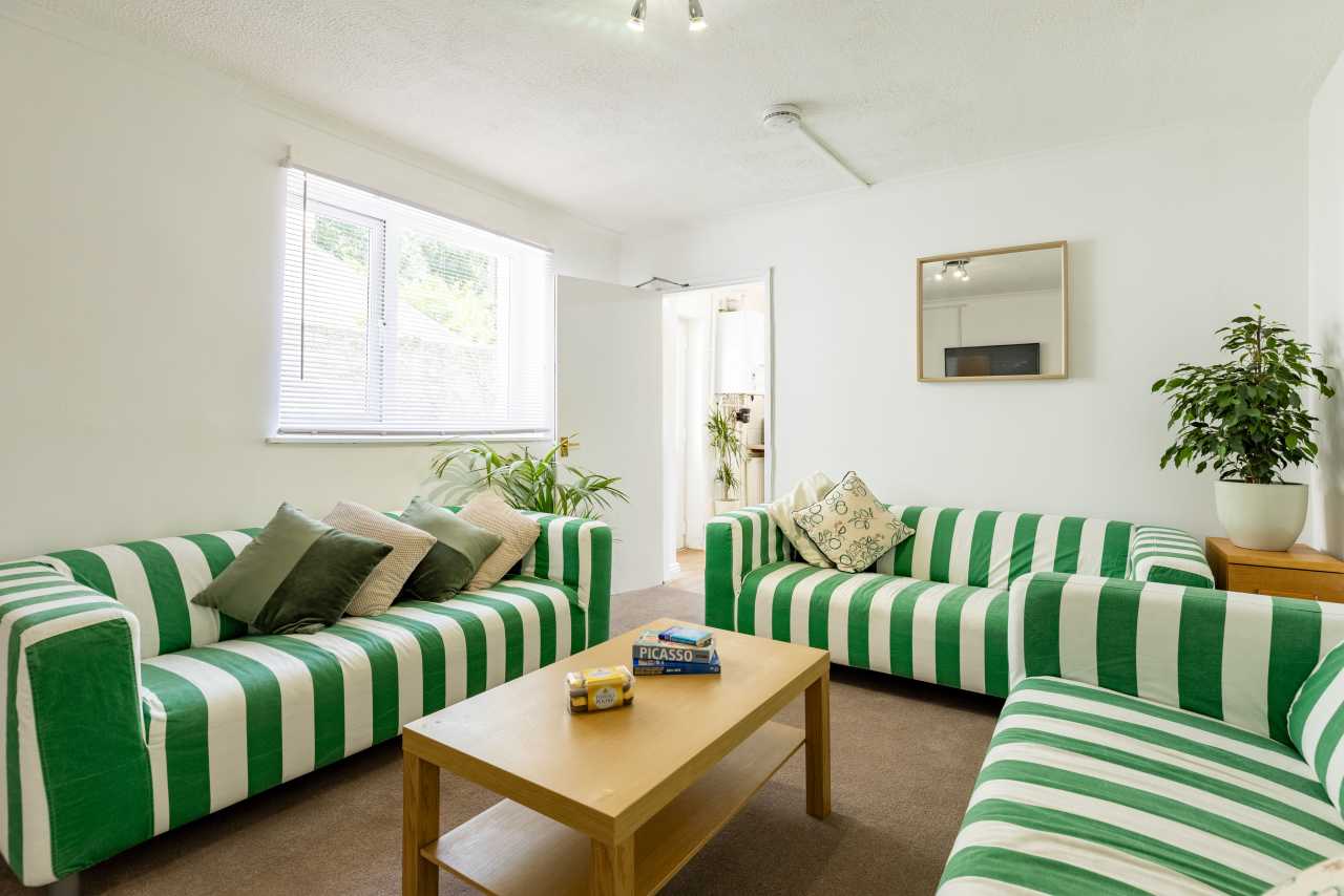 A bright communal lounge with green-and-white striped sofas, a wooden coffee table, a window, and potted plants.