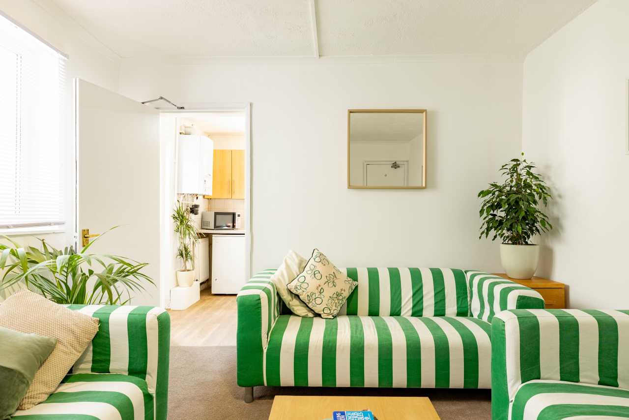Bright living area with green-and-white striped sofas, plants, a wall mirror, and a kitchenette visible through an open doorway.