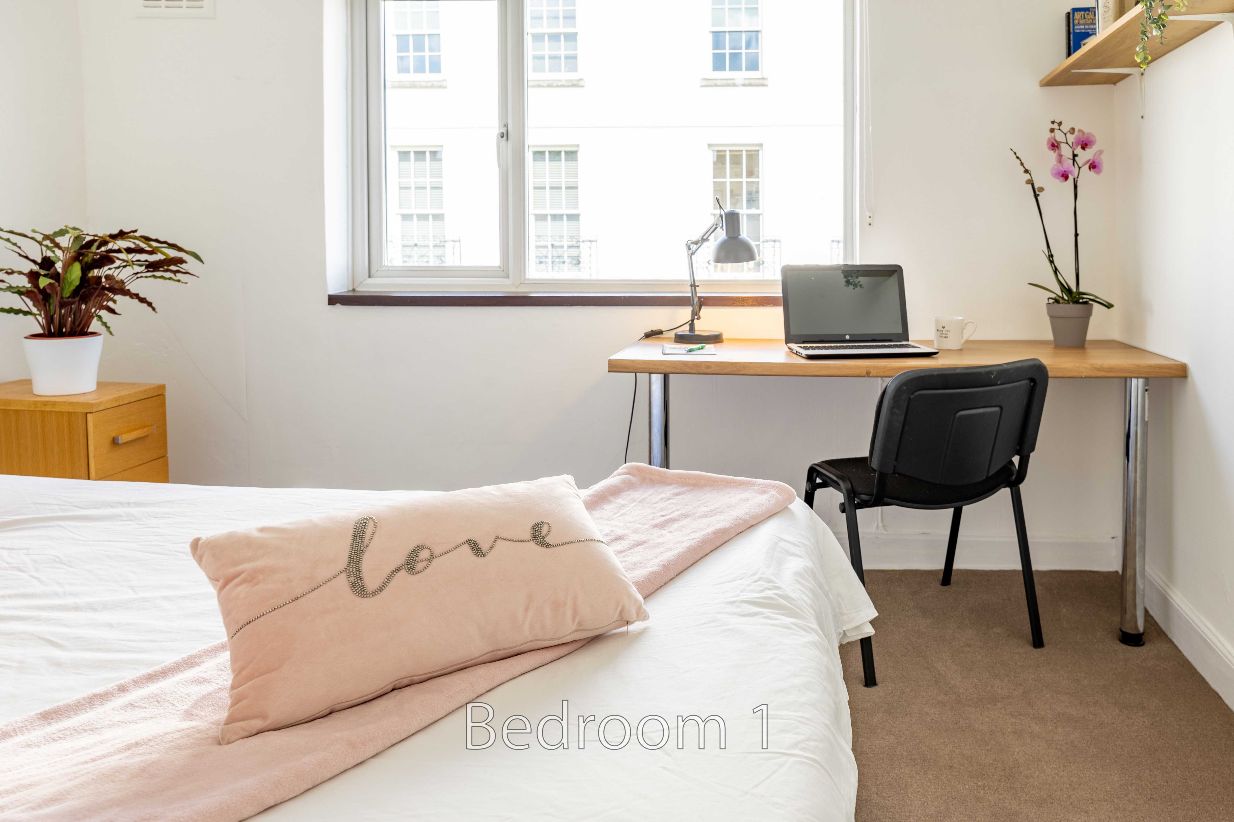 A bedroom with a bed, desk, chair, window, plants and storage furniture.