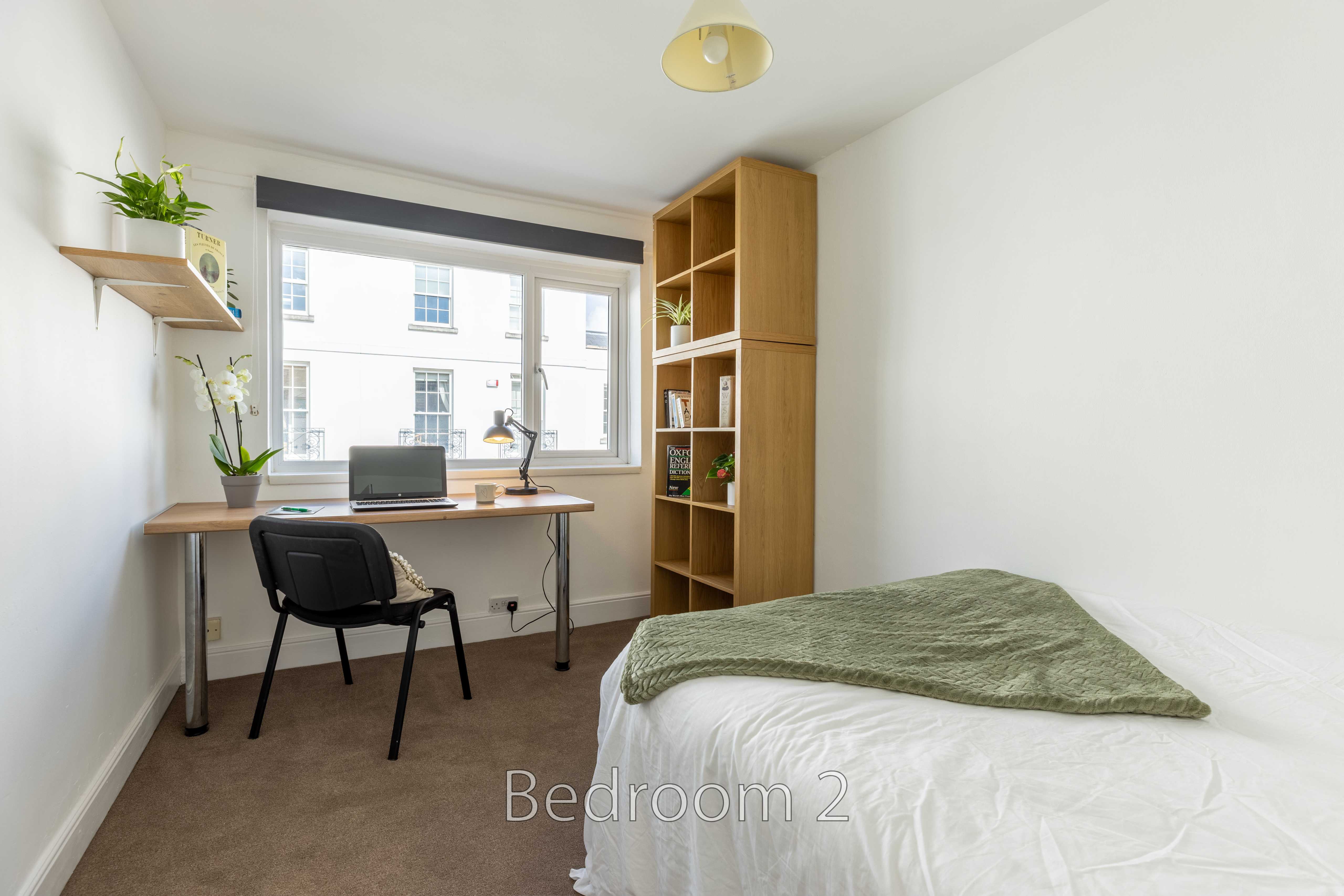A bedroom with a single bed, desk, chair, shelving and a window.
