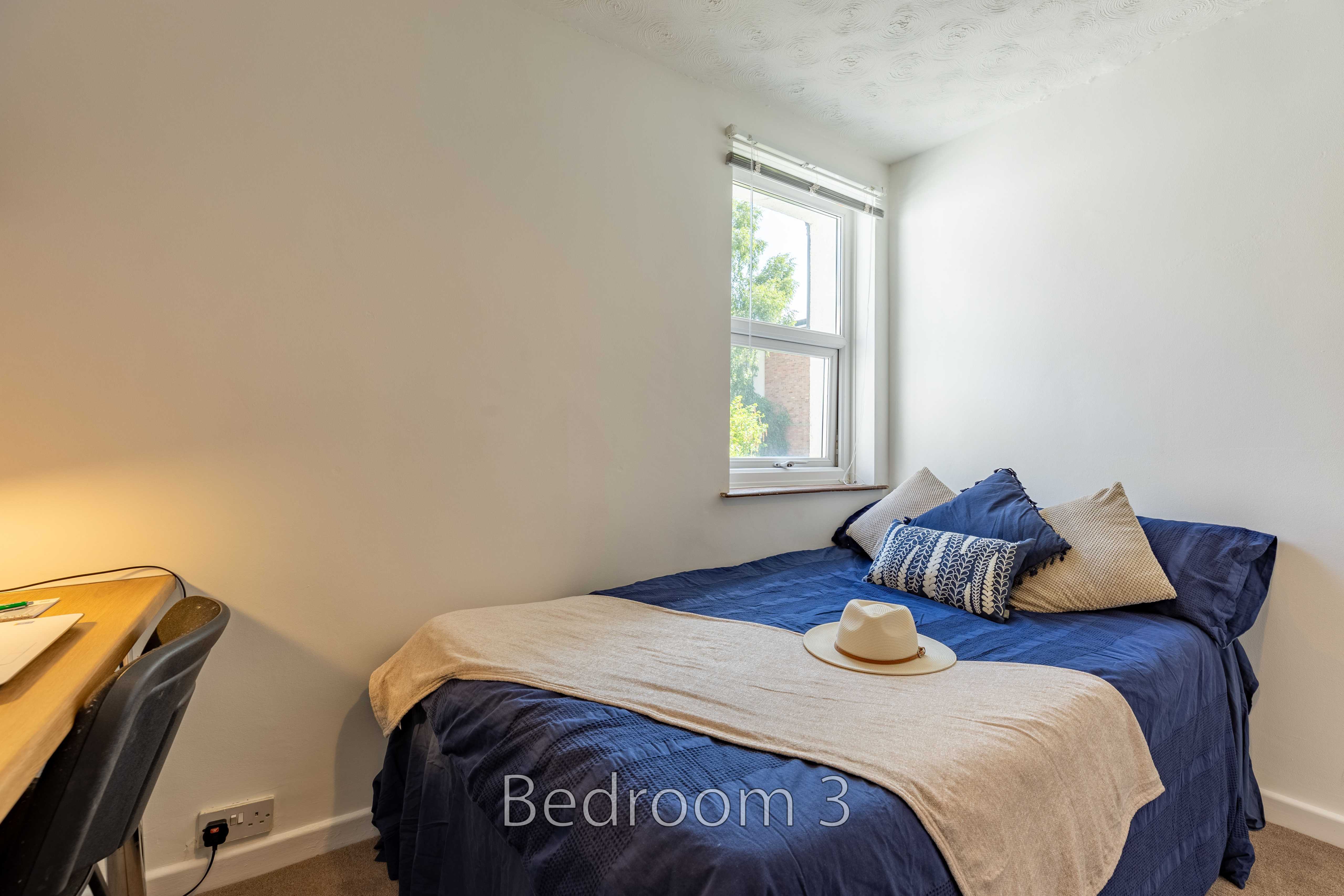 A bedroom with a double bed, blue bedding, pillows, a window, and a desk.